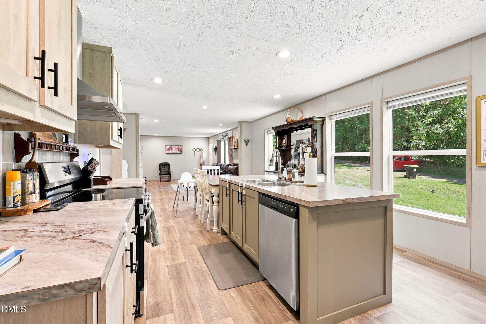 Modern kitchen with light wood cabinets, center island with dishwasher, and large windows overlooking a green yard. Dining table and chairs are visible in the background. Light wood flooring throughout.