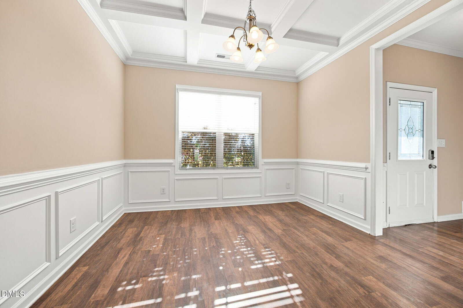 Empty room with beige walls, white wainscoting, wood-style flooring, a window with blinds, ceiling lights, and a white door with a glass panel. Sunlight creates patterns on the floor.