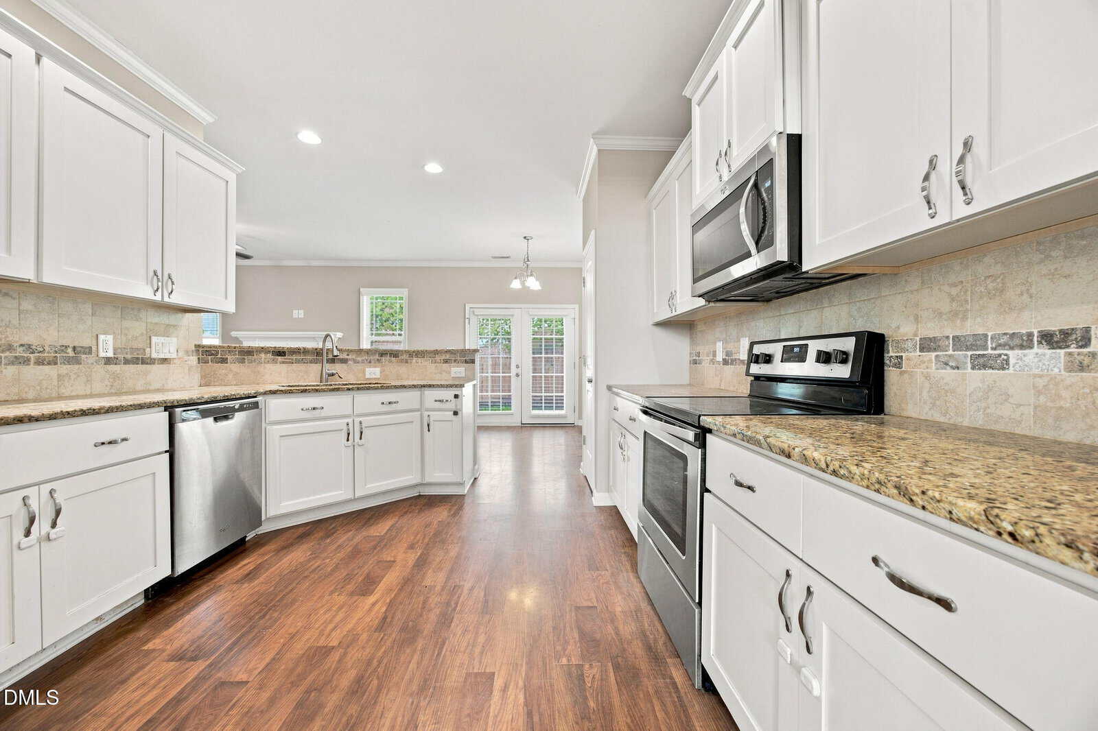 Modern kitchen with white cabinets, granite countertops, stainless steel appliances, tile backsplash, and wood flooring. The room is well-lit with recessed lighting and natural light from glass doors in the background.