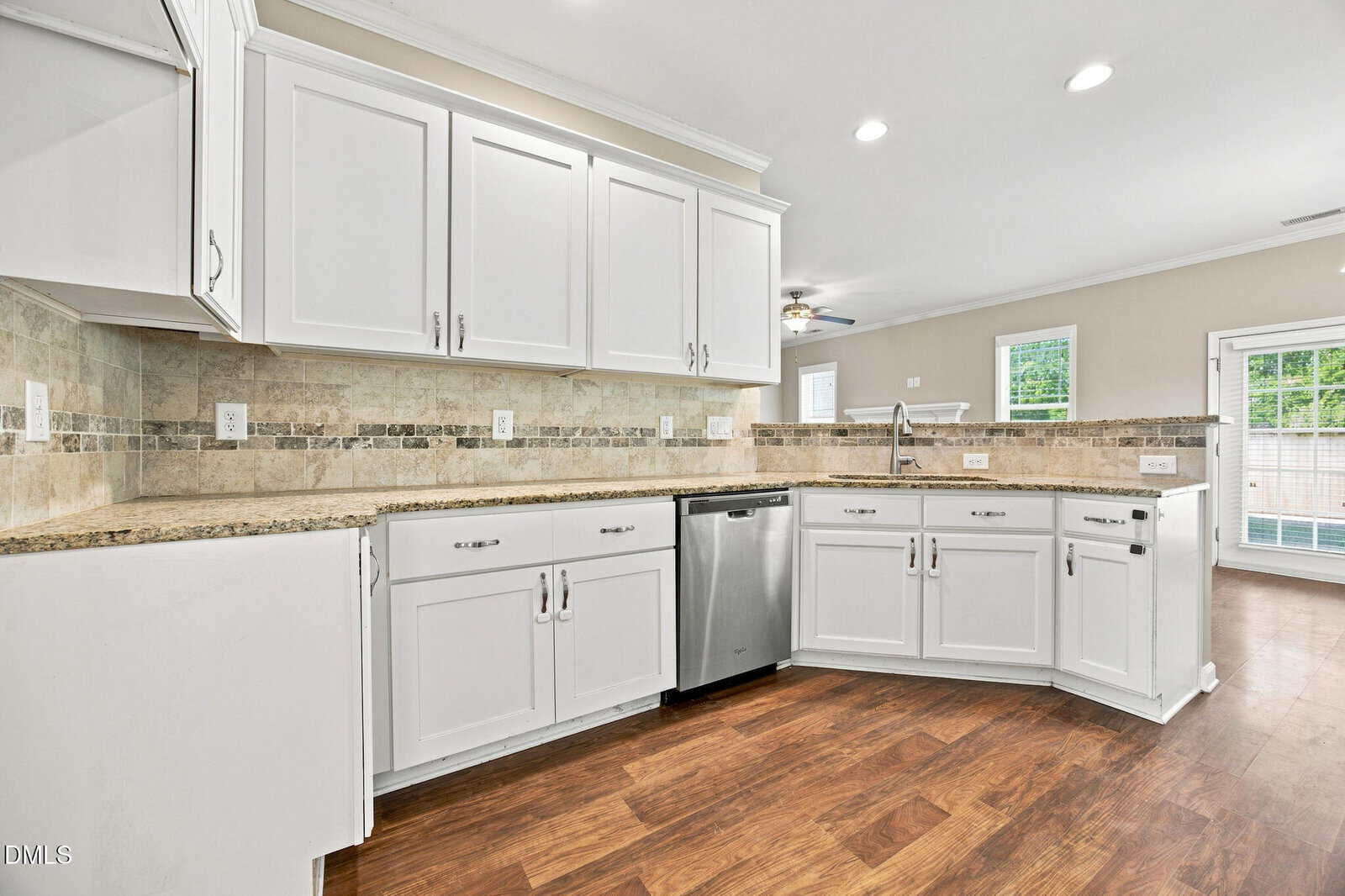 Modern kitchen with white cabinets, granite countertops, tile backsplash, stainless steel dishwasher, and wooden floors. The space opens into a living area with large windows and a door leading outside.