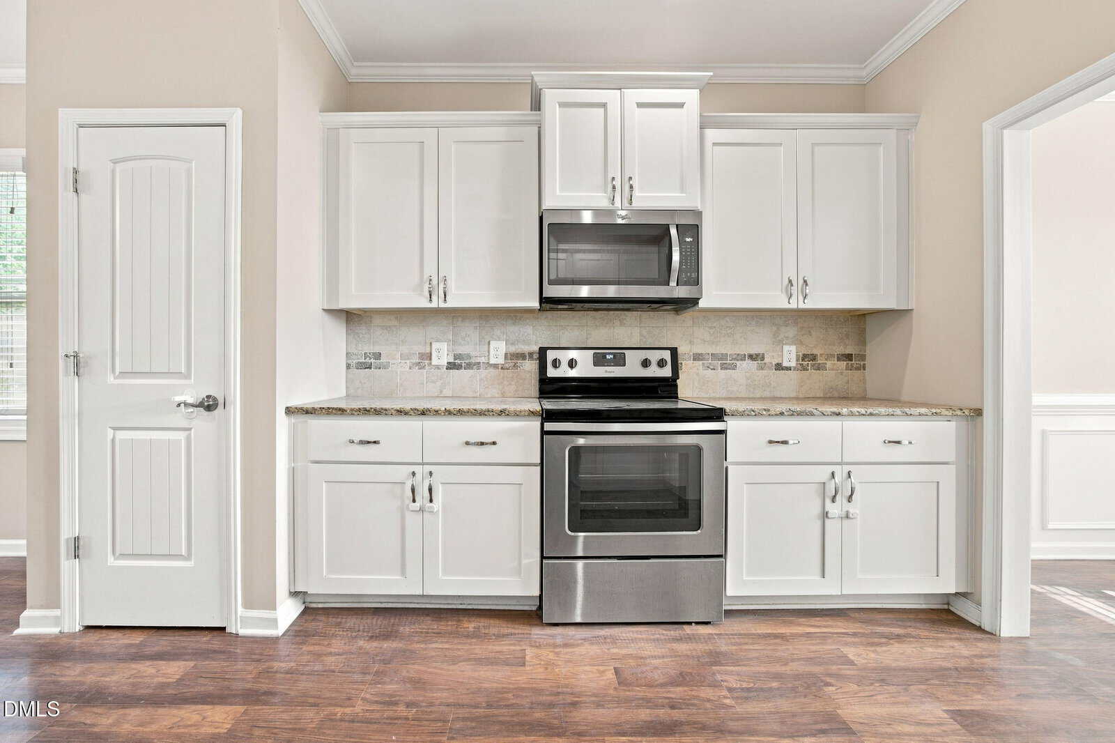 A modern kitchen features white cabinets, a stainless steel oven with a matching microwave above, granite countertops, light tile backsplash, and dark wood floors. A closed white door and a window are on the left.