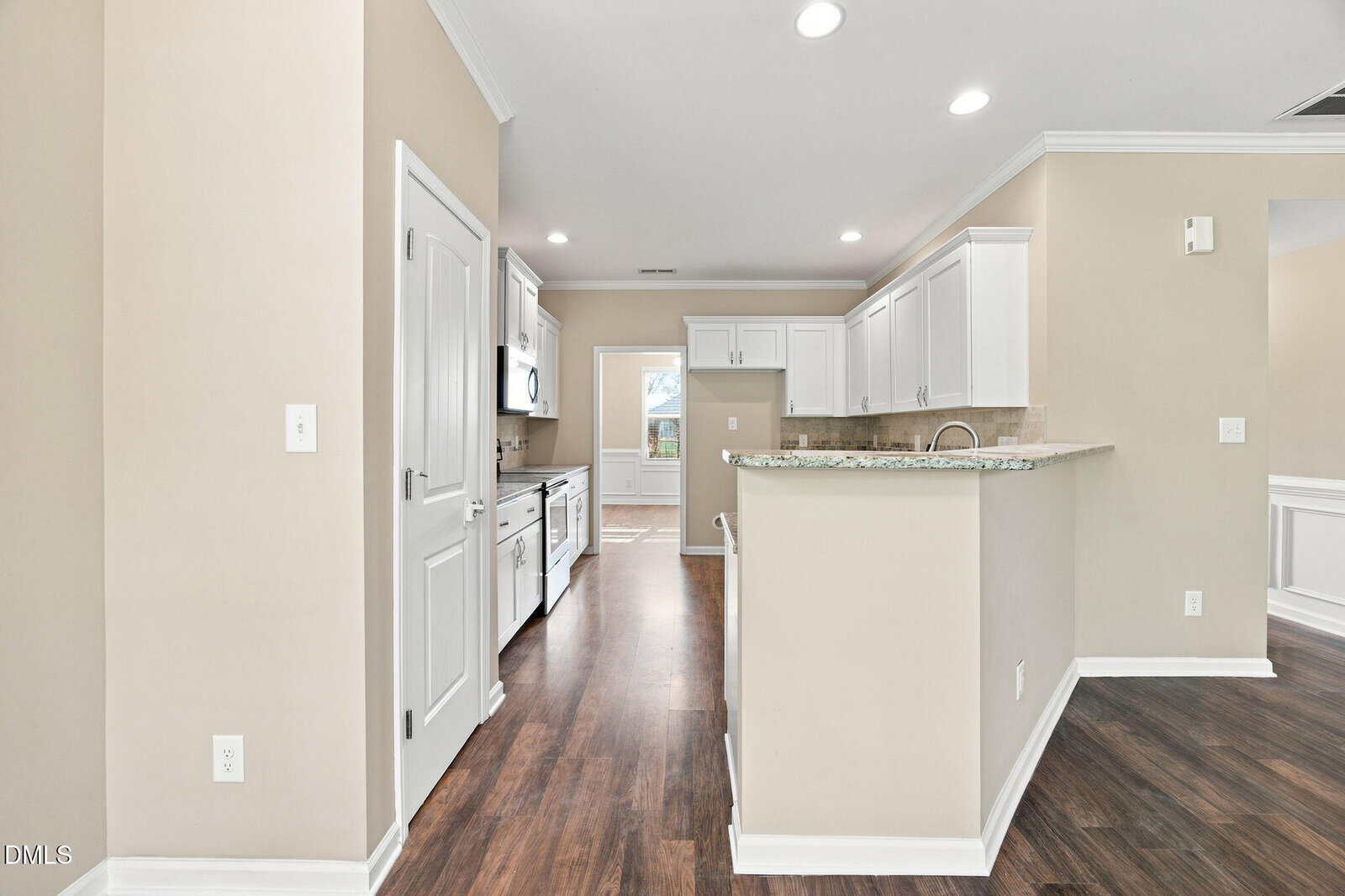 Modern kitchen with white cabinets, stainless steel appliances, granite countertops, and dark wood floors. There is a breakfast bar and an open doorway leading to a dining area with natural light. Walls are painted a light beige.