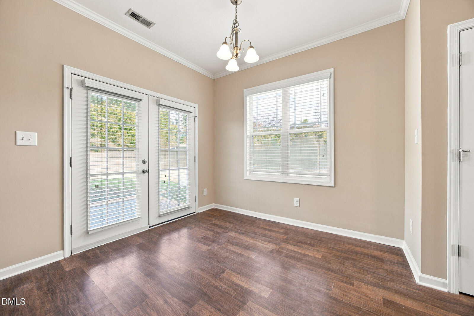 Bright, empty room with tan walls, white trim, wood flooring, a window with blinds, and glass double doors leading outside. Ceiling light fixture with three bulbs hangs above.