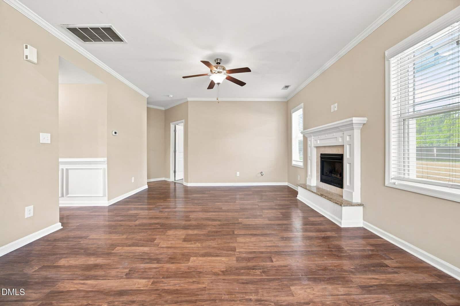 Empty living room with wood flooring, beige walls, white trim, ceiling fan, windows with blinds, and a white fireplace with a granite hearth. A doorway and open entry to another room are visible.