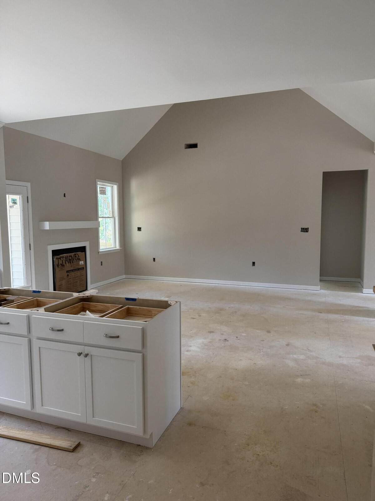 Unfinished room with bare floors, white walls, and vaulted ceiling. White kitchen island cabinets are installed. Fireplace is set into the wall on the left, with nearby windows and a door. Room lacks flooring and fixtures.