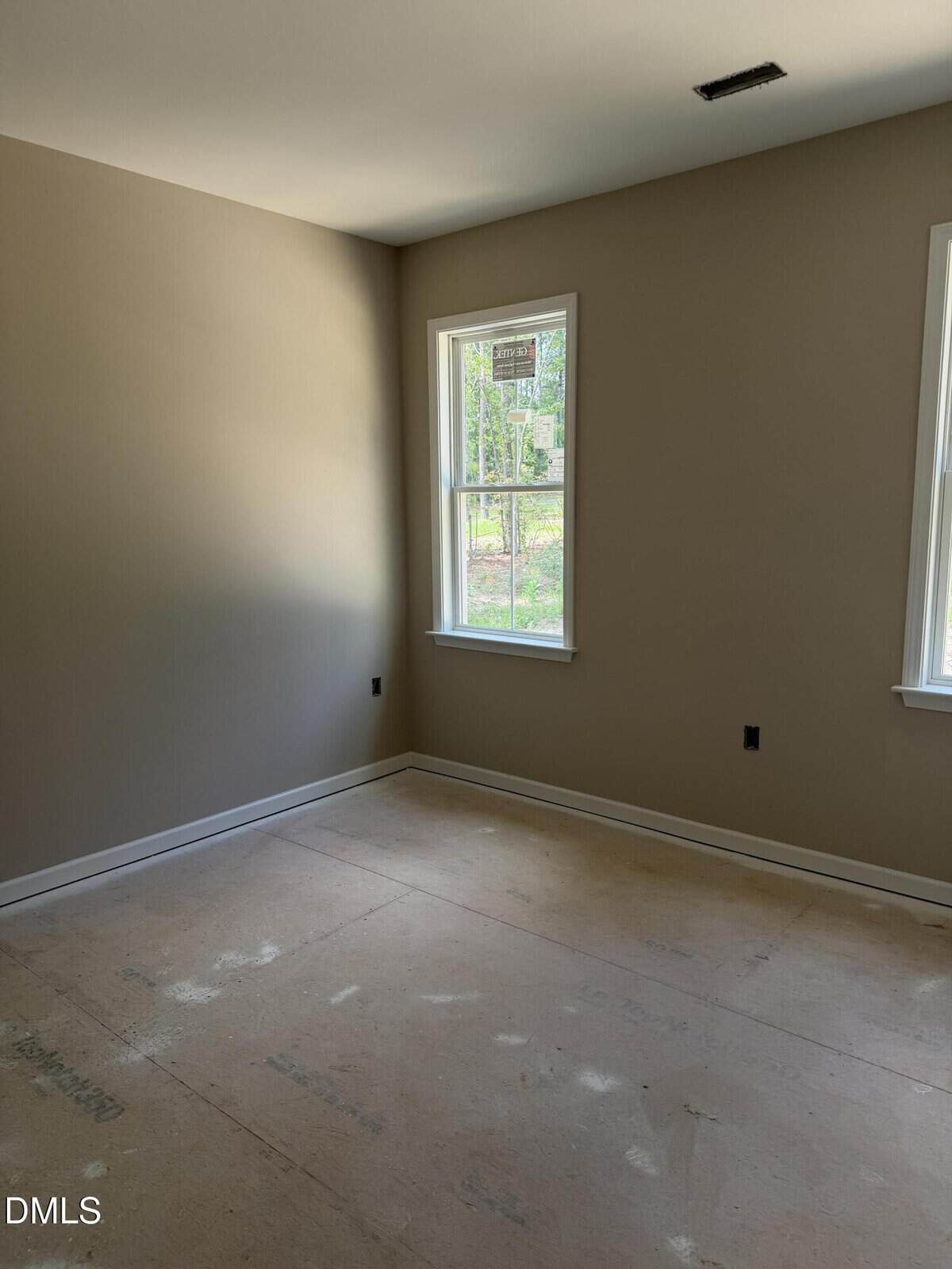 An empty room with beige walls, two windows letting in natural light, and an unfinished floor. No furniture or decorations are present.