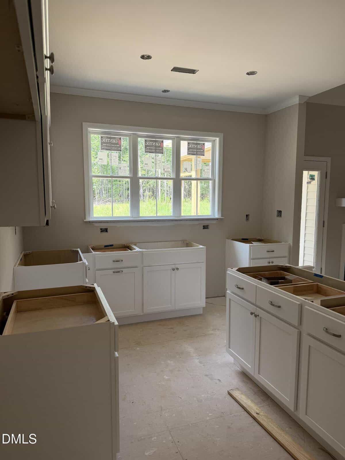 A kitchen under construction with white cabinets installed, but no countertops or appliances yet. Large windows let in natural light, and cabinets are arranged along the walls and in the center.