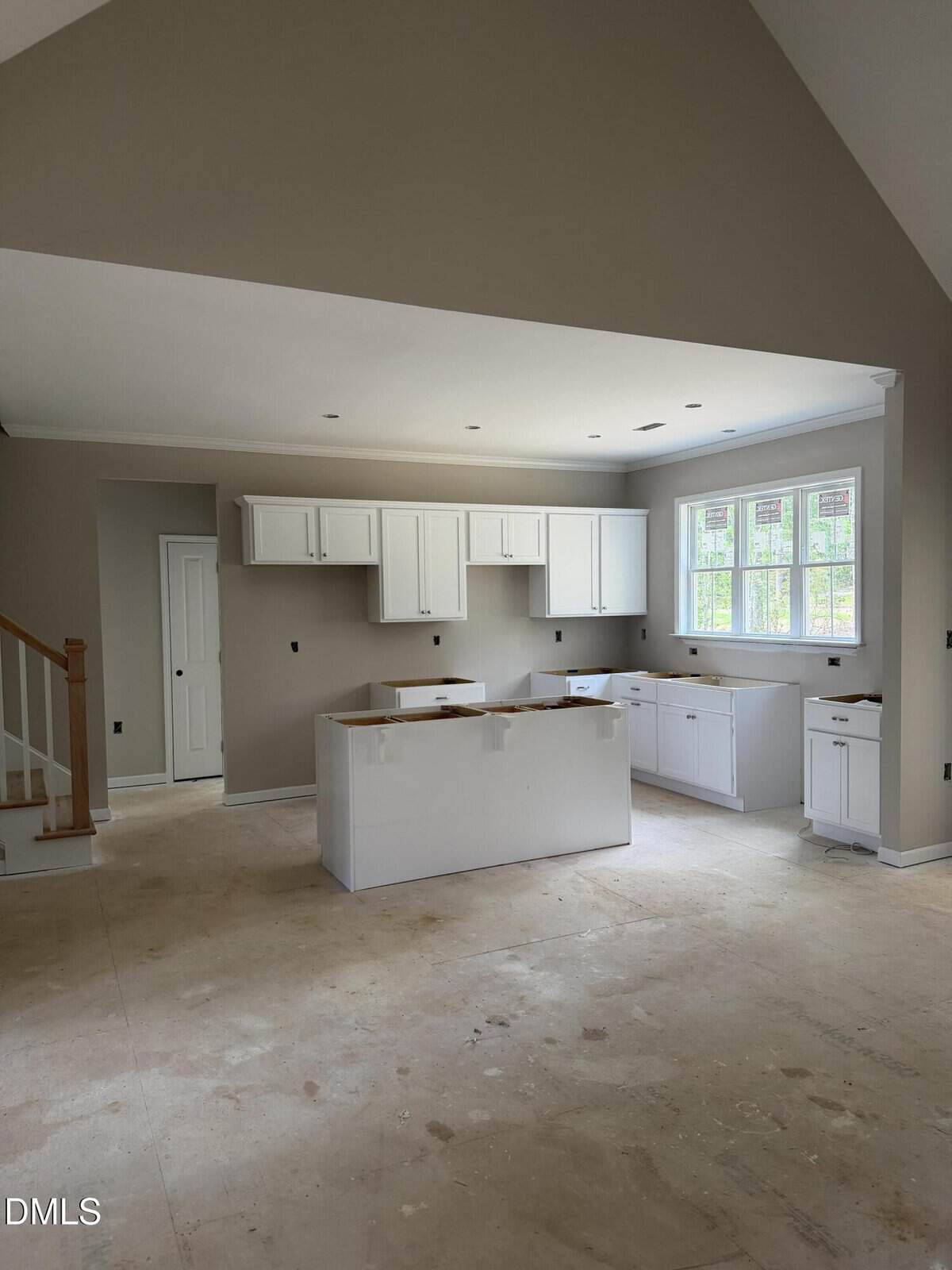 A kitchen under construction with unfinished floors, white cabinets, a center island, and large windows. The walls are painted beige, and there are no appliances or countertops installed yet. Stairs are visible on the left.