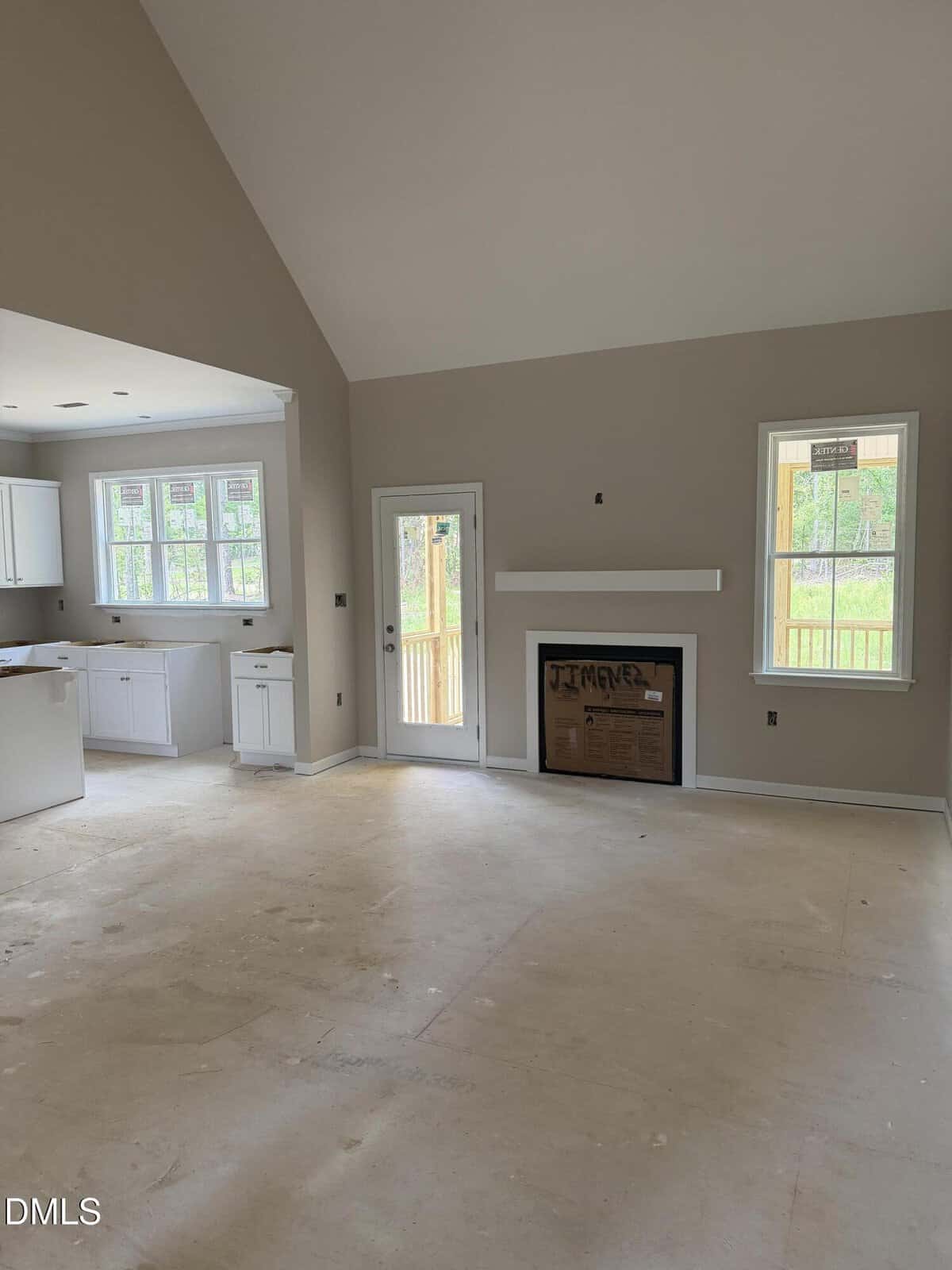 Unfurnished living room with beige walls, a fireplace covered in cardboard, a mounted shelf above, and large windows. The kitchen with white cabinets is partially visible on the left. Unfinished floor.