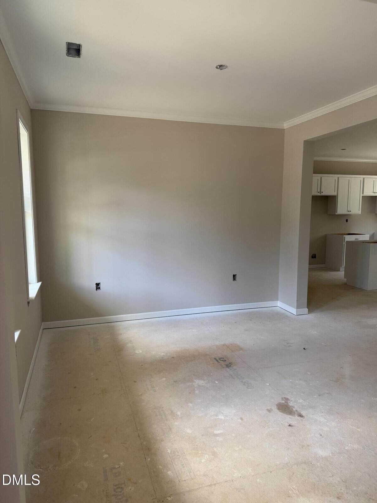 Empty, unfinished room with beige walls, white trim, and bare floor. An adjacent room with white cabinets is visible through an open doorway. Sunlight comes through a window on the left wall.