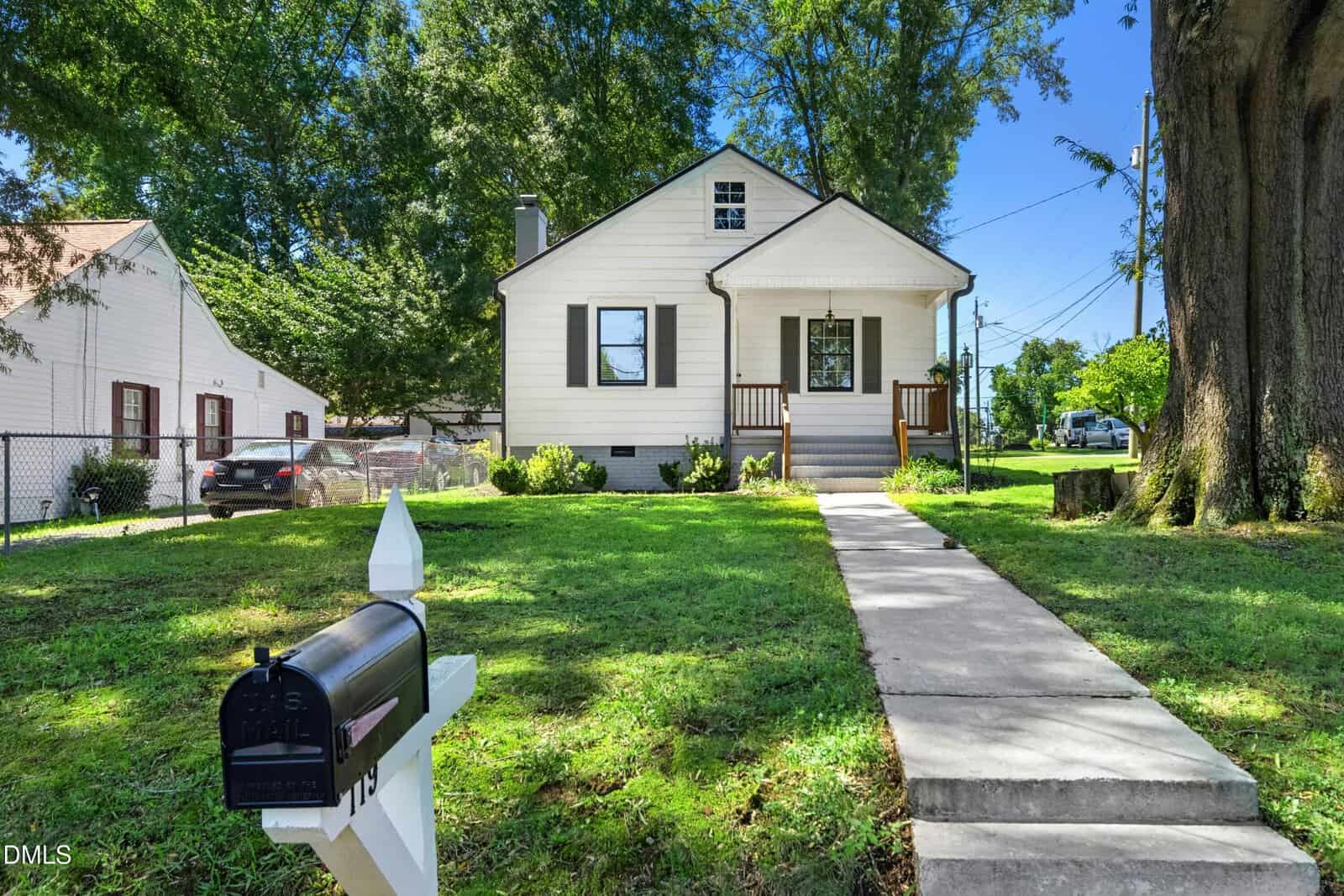 A small cream-colored house with dark trim, a front porch, stairs, and a paved walkway leading to a black mailbox. The yard is grassy with large trees, and a car is visible parked beside a neighboring house.
