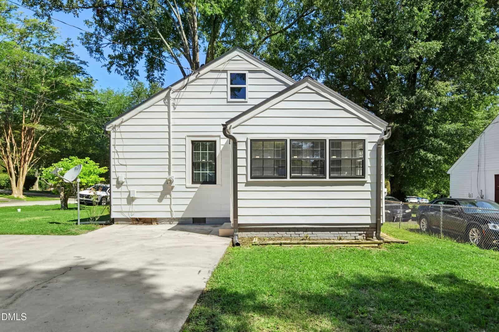 A small, white house with horizontal siding, black-framed windows, and a concrete driveway, surrounded by green grass and tall trees on a sunny day.