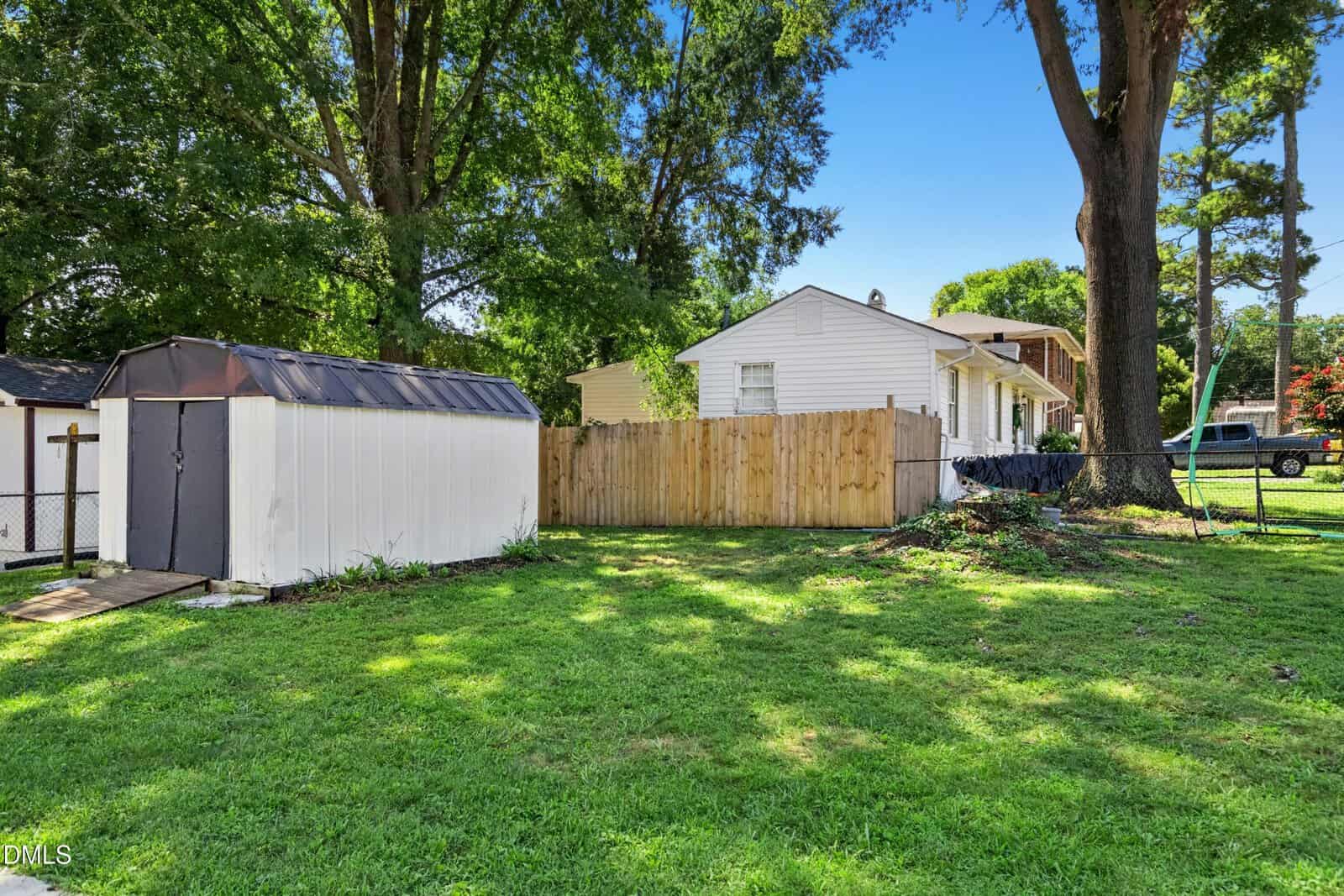A fenced backyard with green grass, a white house, large trees, a small white and black shed, and a hammock. A pickup truck is visible in the background near a driveway.