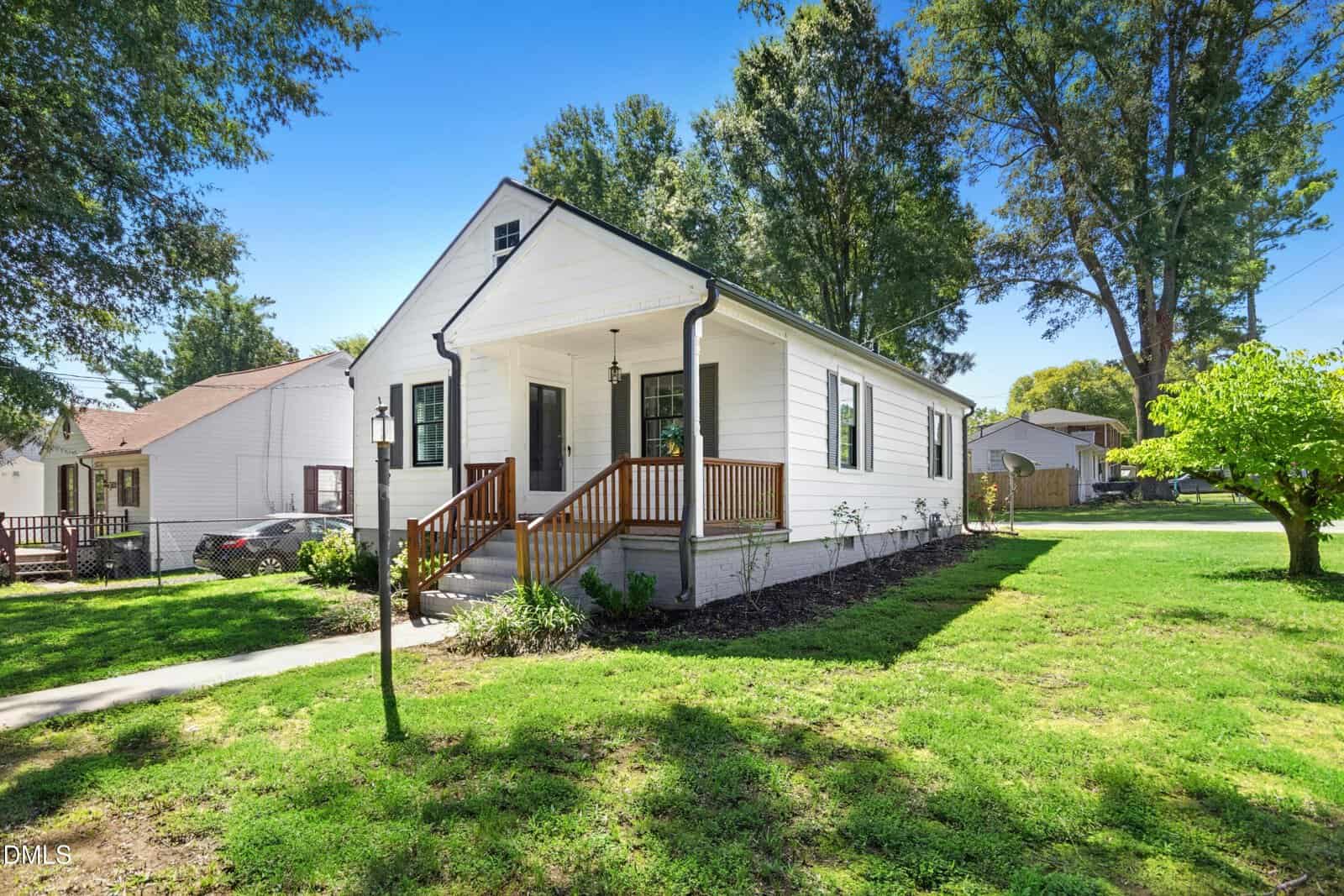 A small white single-story house with a covered front porch, green shutters, and a manicured lawn sits on a sunny day with trees and neighboring houses visible in the background.