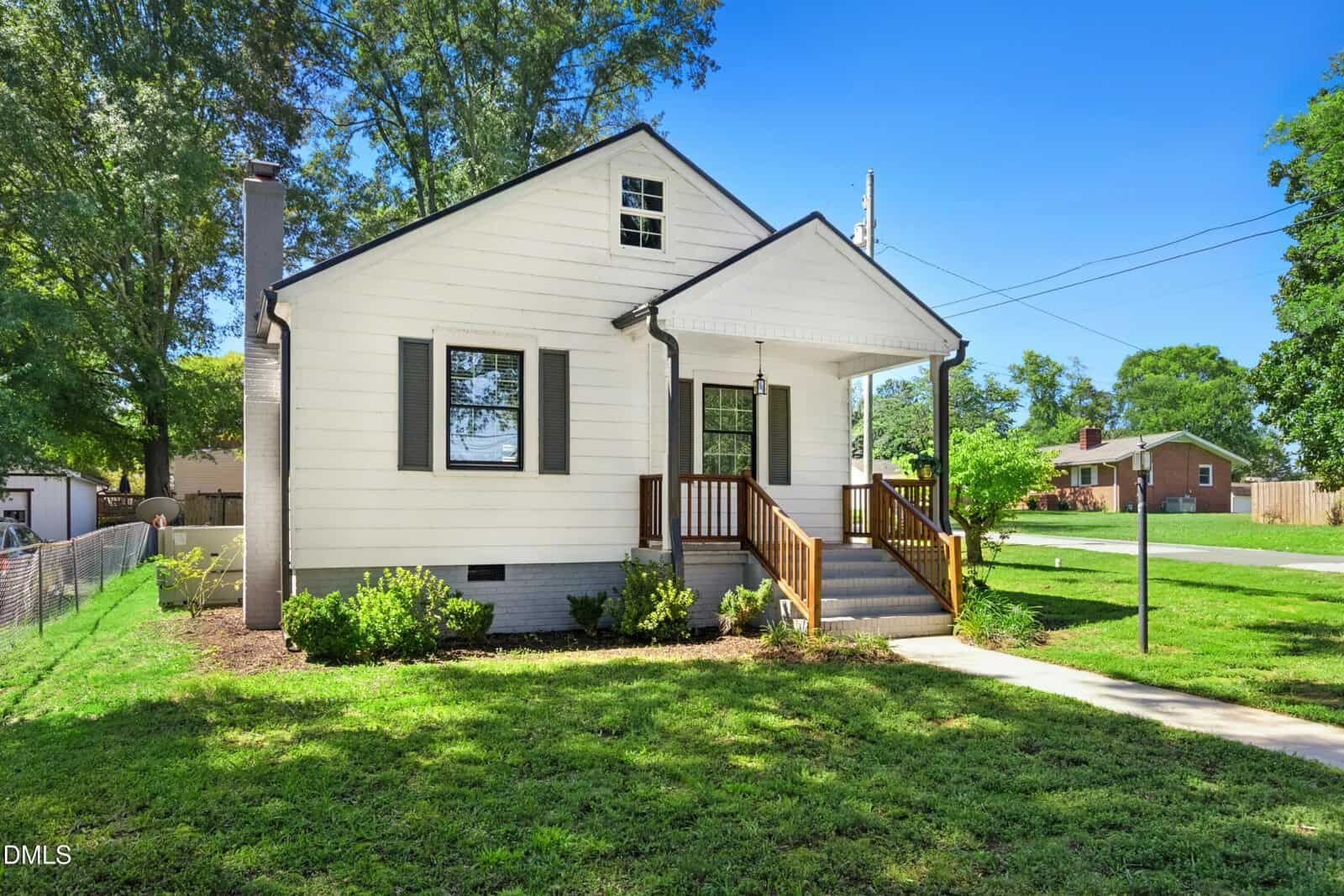 A white single-story house with a covered front porch, dark shutters, and stairs leading to the entrance. The house sits on a grassy lawn with bushes and trees, and a sidewalk runs along the front.