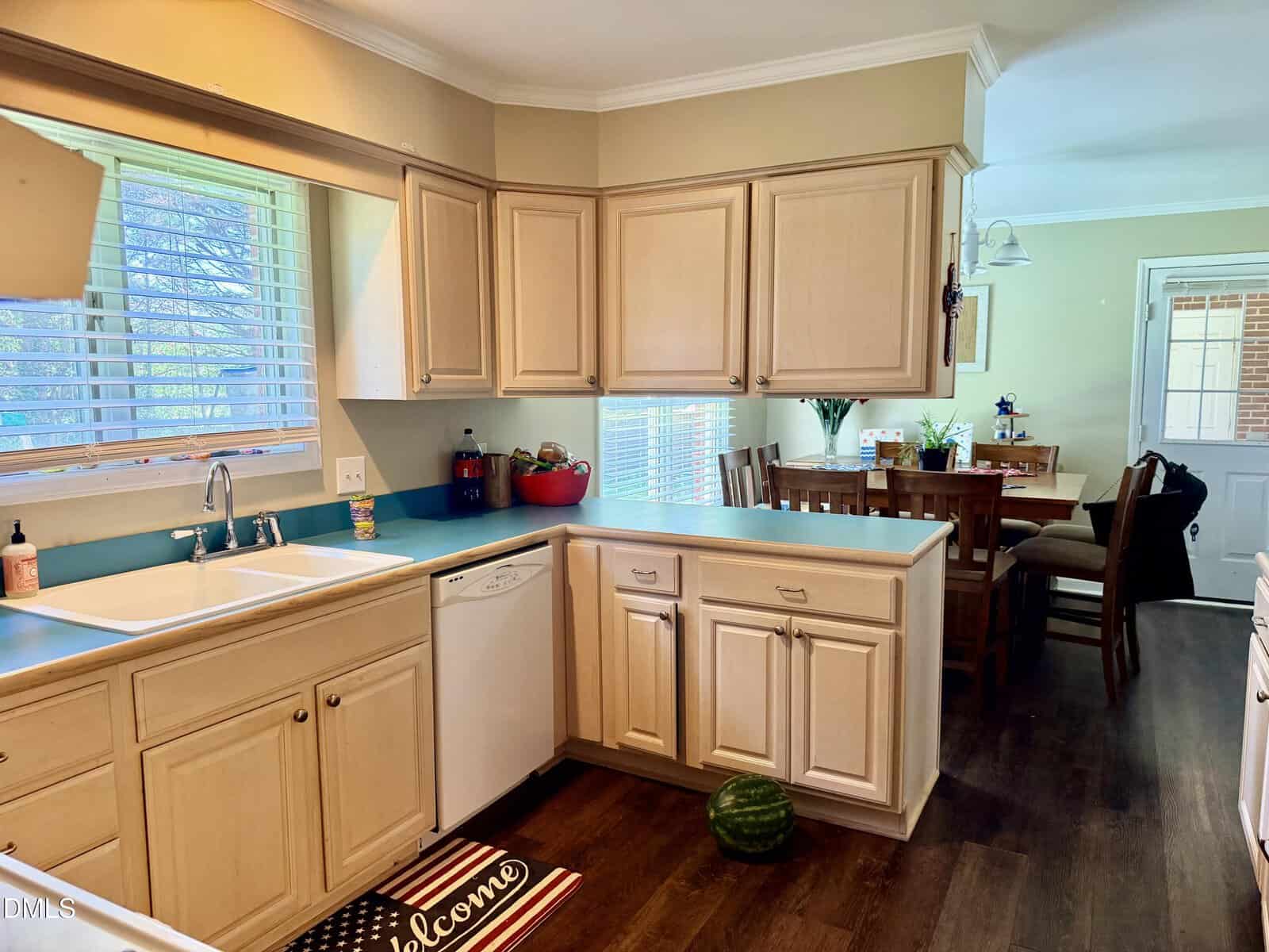 A kitchen with light wood cabinets, a blue countertop, a white sink, and a dishwasher. A watermelon sits on the floor. The kitchen opens to a dining area with a wooden table and chairs by a window.