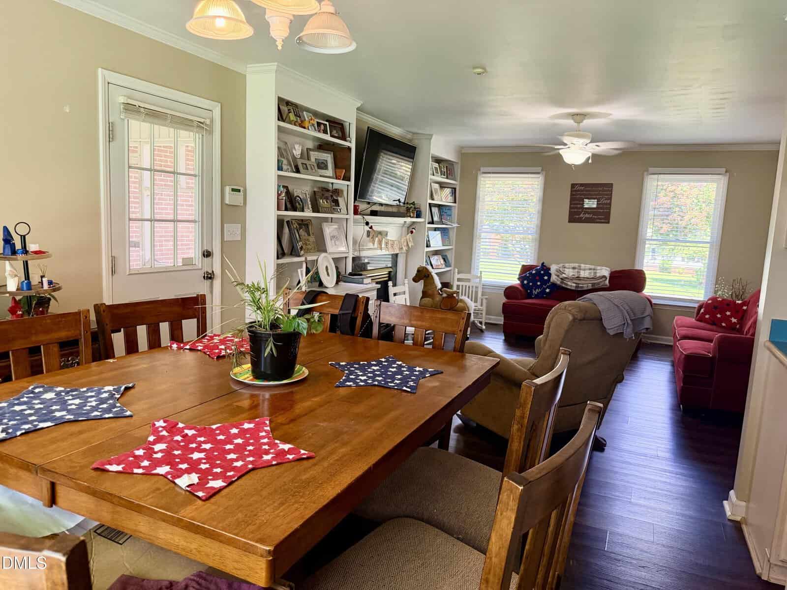 A dining area with a wooden table and chairs is in the foreground. The living area in the background has armchairs, a TV, bookshelves, large windows, and decor. There are patriotic star placemats on the table.