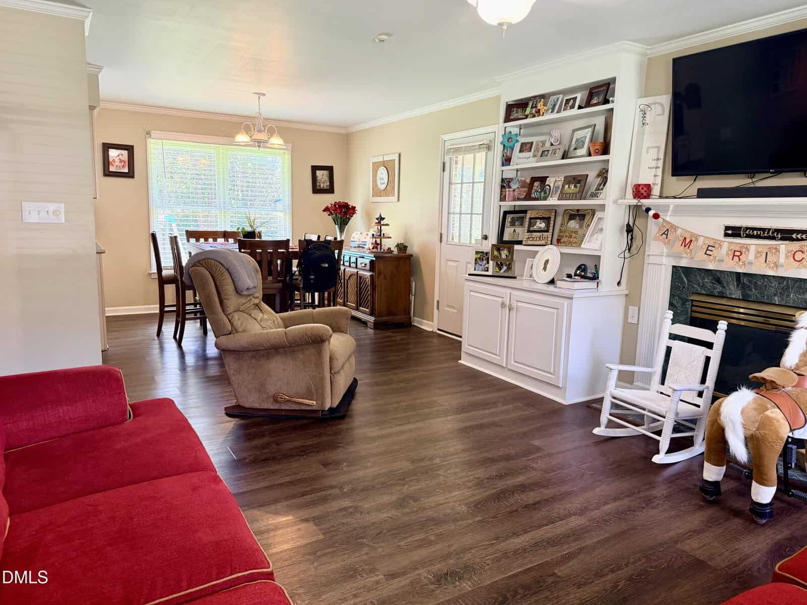 A living room with wood floors, a red couch, tan recliner, white shelving, and a fireplace. A dining area with a table and chairs is in the background near large windows. Decorations and framed photos are displayed throughout.