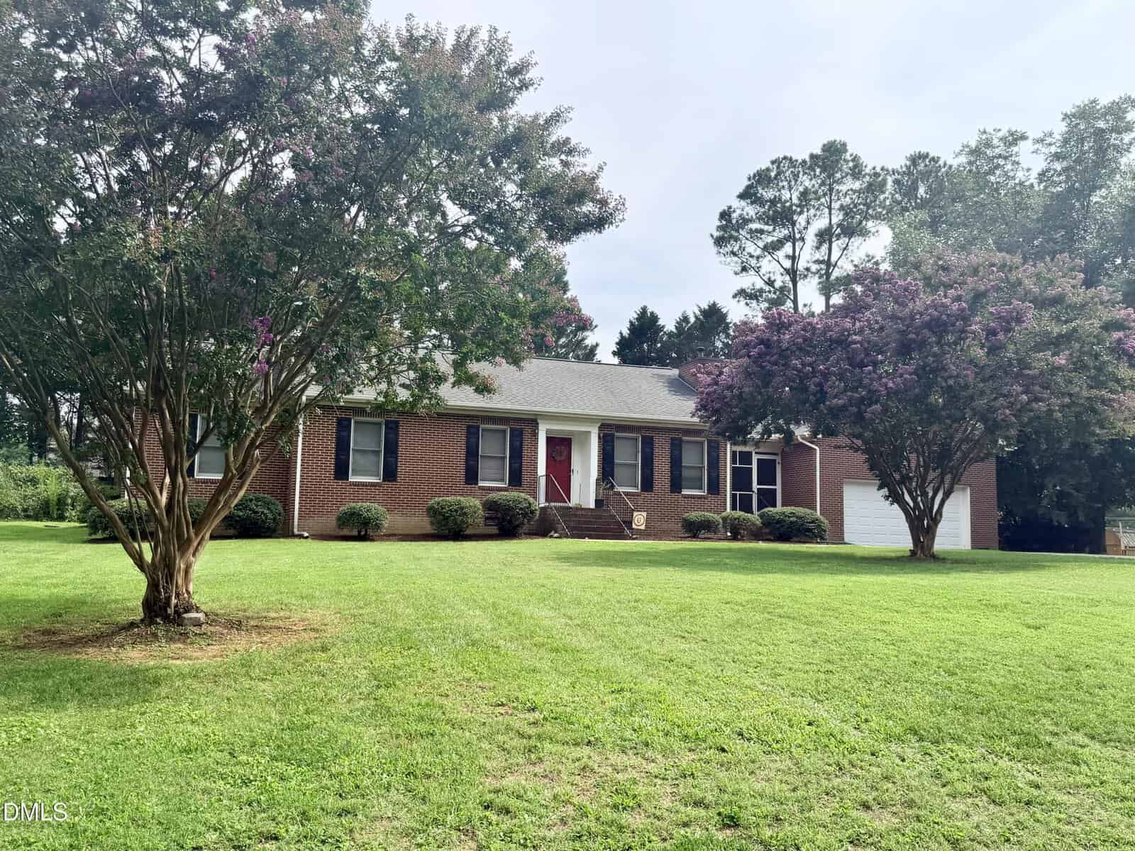 A one-story brick house with a red front door, black shutters, a two-car garage, and a large, well-kept lawn. Two flowering trees and several bushes are in the front yard. The sky is partly cloudy.