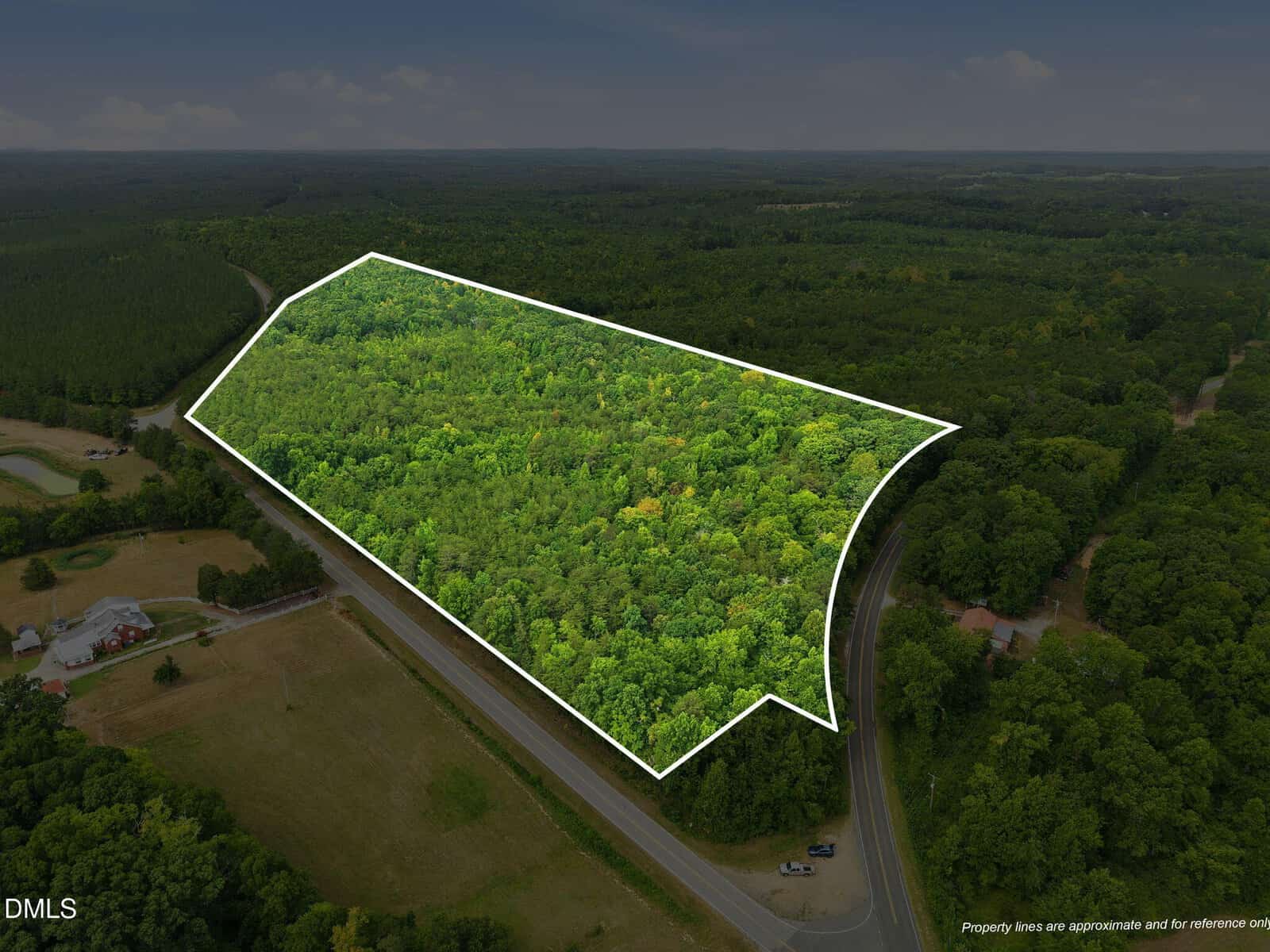 Aerial view of a large, densely wooded plot of land outlined in white, surrounded by roads and some cleared areas with buildings, with a forested landscape in the background.