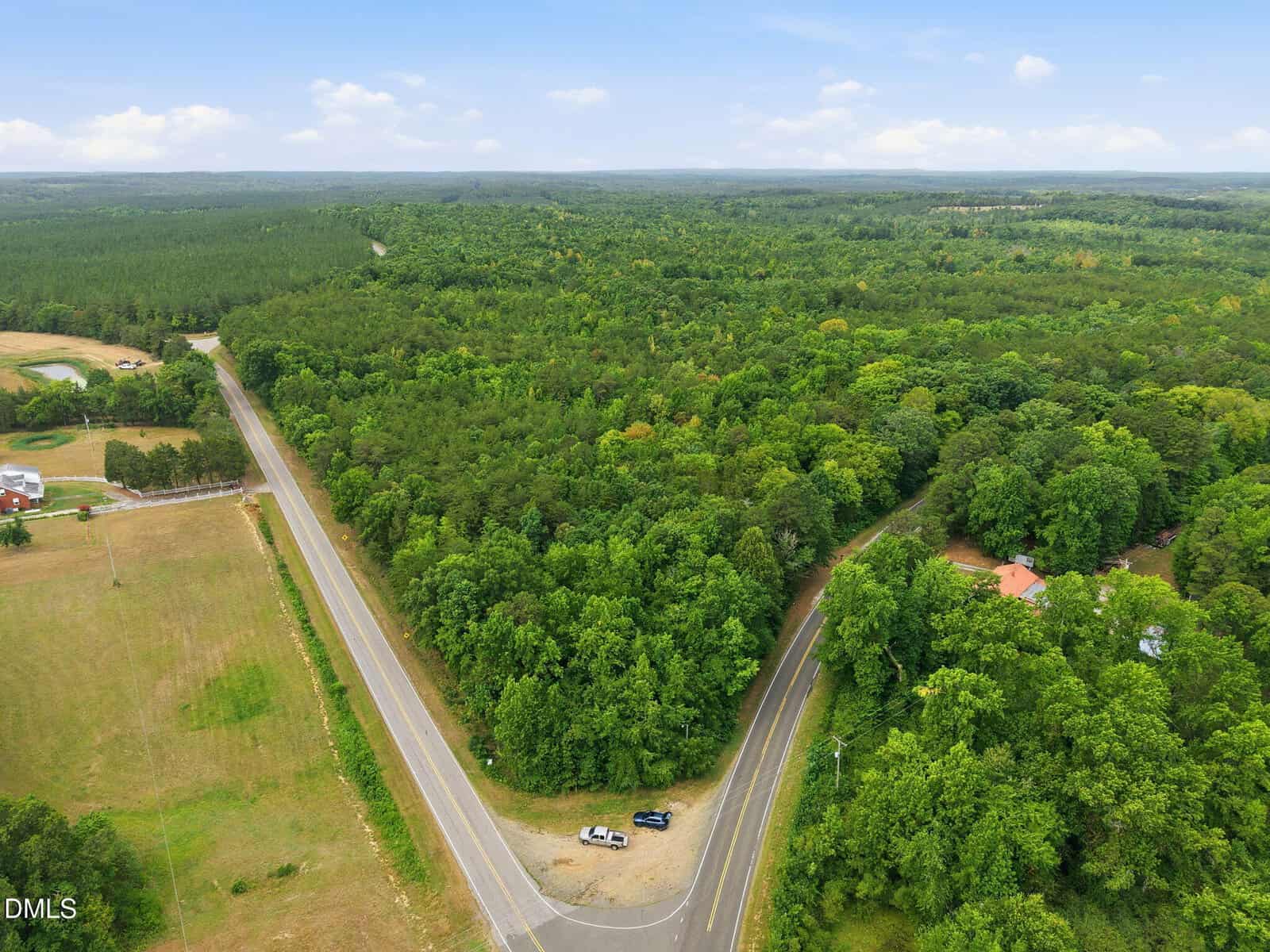 Aerial view of a forested area bordered by a curved road and a straight road intersection, with a few parked vehicles and buildings partially visible among the trees. The sky is mostly clear with scattered clouds.