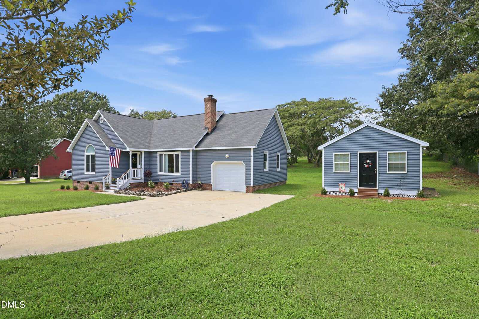 A light blue house with a front porch and attached chimney sits beside a matching detached garage, both with white trim, at 147 Bridle Trail in Youngsville. The property features a large driveway, green lawn, and trees under a blue sky.