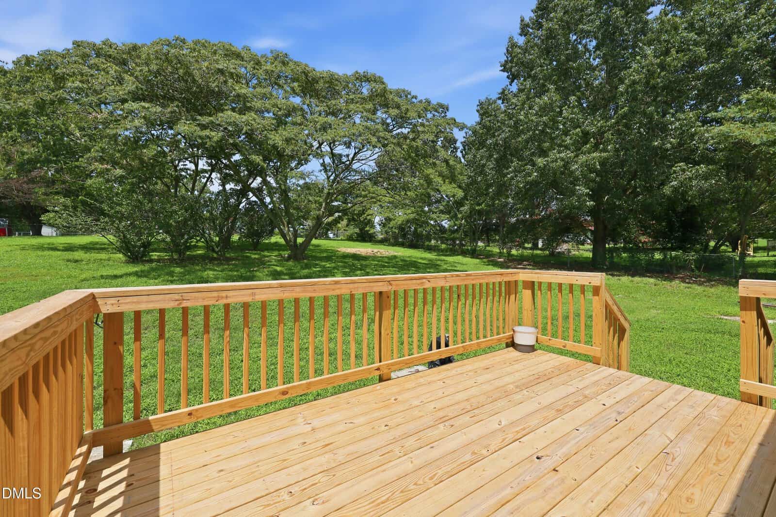 A wooden deck with a railing overlooks a grassy yard and several large trees under a clear blue sky. A small white container sits in the corner of the deck.