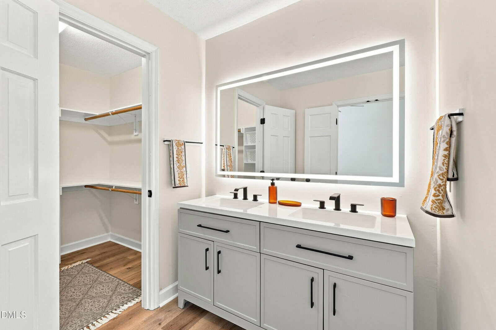 Modern bathroom with a double-sink vanity, black fixtures, large backlit mirror, two towels, amber soap dispensers, white walls, and a view into a walk-in closet with wooden shelves and hanging rods.