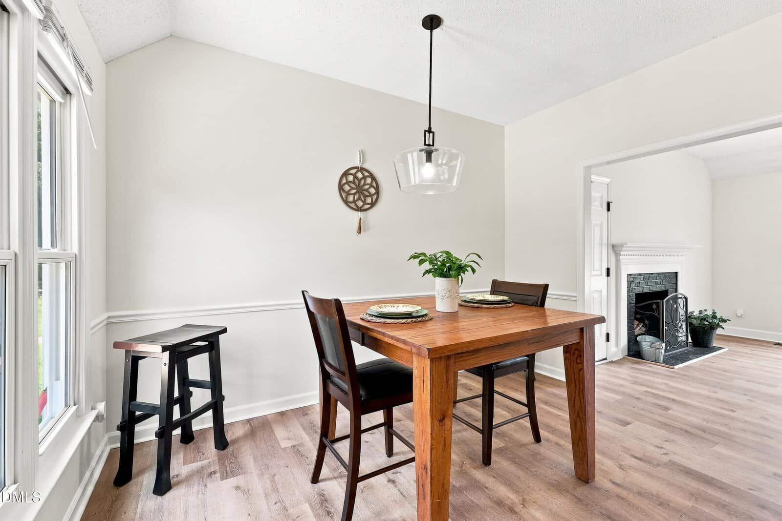 A small dining area with a wooden table, two chairs, a bench, a potted plant centerpiece, a wall decoration, pendant light, and large windows. The area opens to a living room with a fireplace.