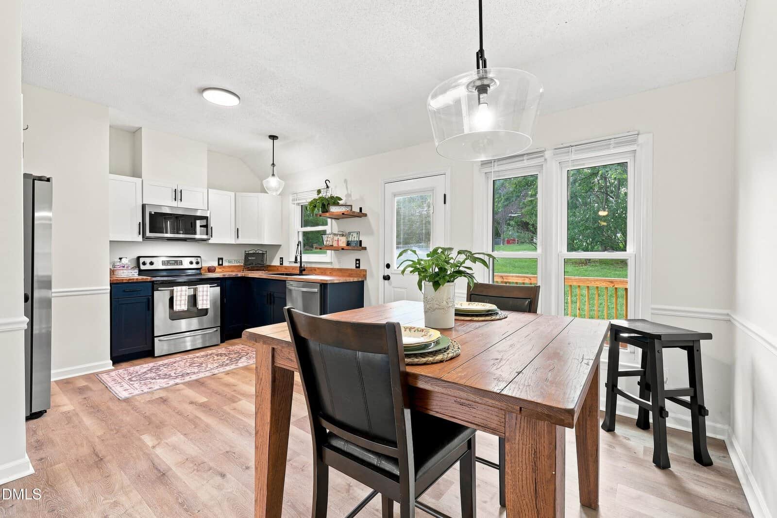 Bright kitchen with white cabinets, stainless appliances, and wood countertops. A wooden dining table with black chairs sits by large windows overlooking a green backyard. Pendant lights hang over the table and kitchen.