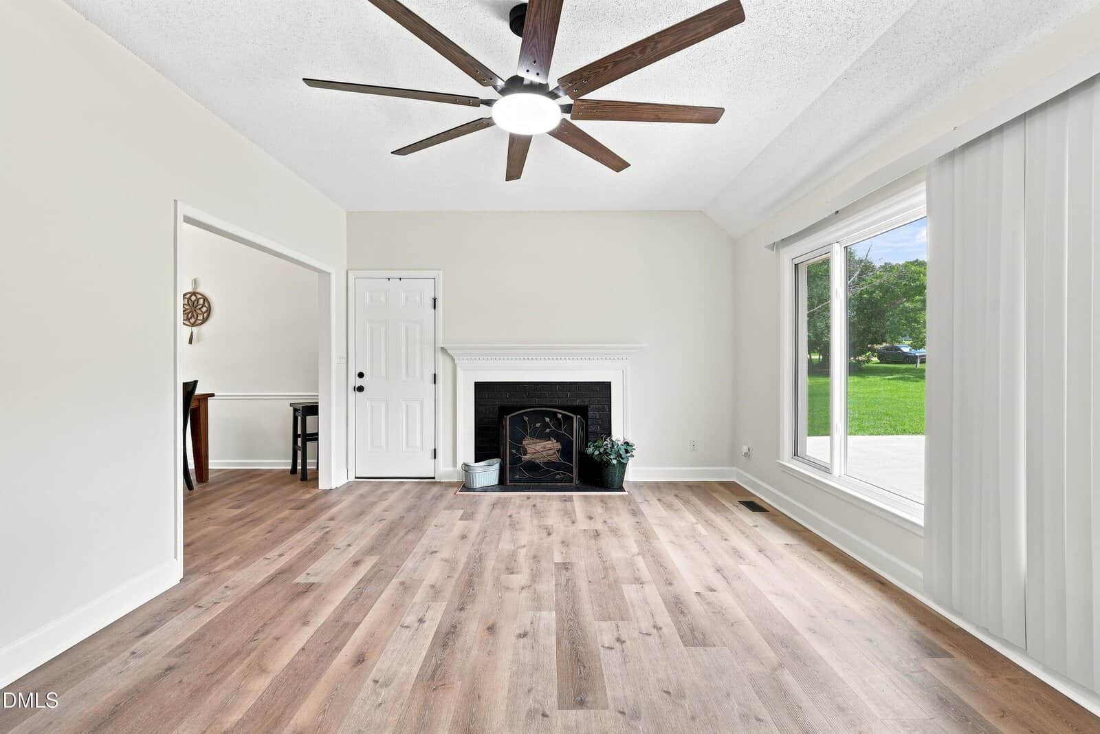 Bright living room with wood floors, white walls, large ceiling fan, fireplace, potted plant, and a sliding glass door leading to a green lawn outside. Dining area visible through an open doorway on the left.