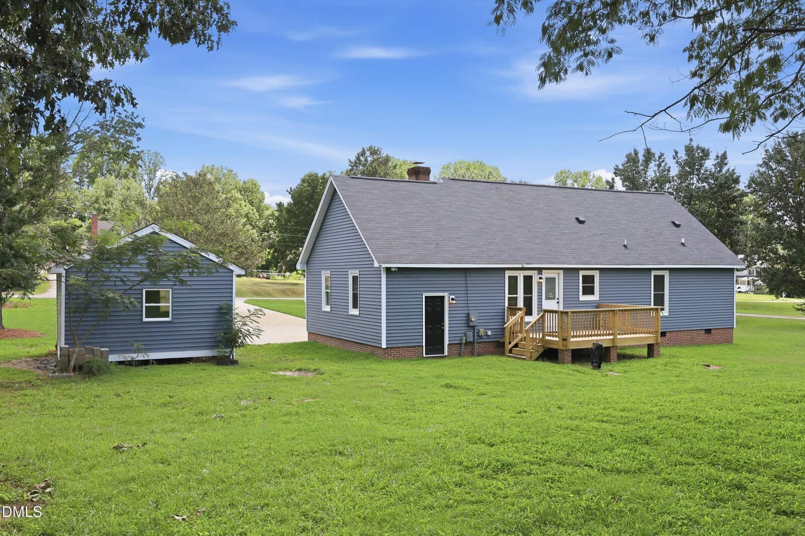 A blue house with a small wooden deck, large grassy yard, and a matching blue shed. Trees surround the property, and the sky is bright with scattered clouds.