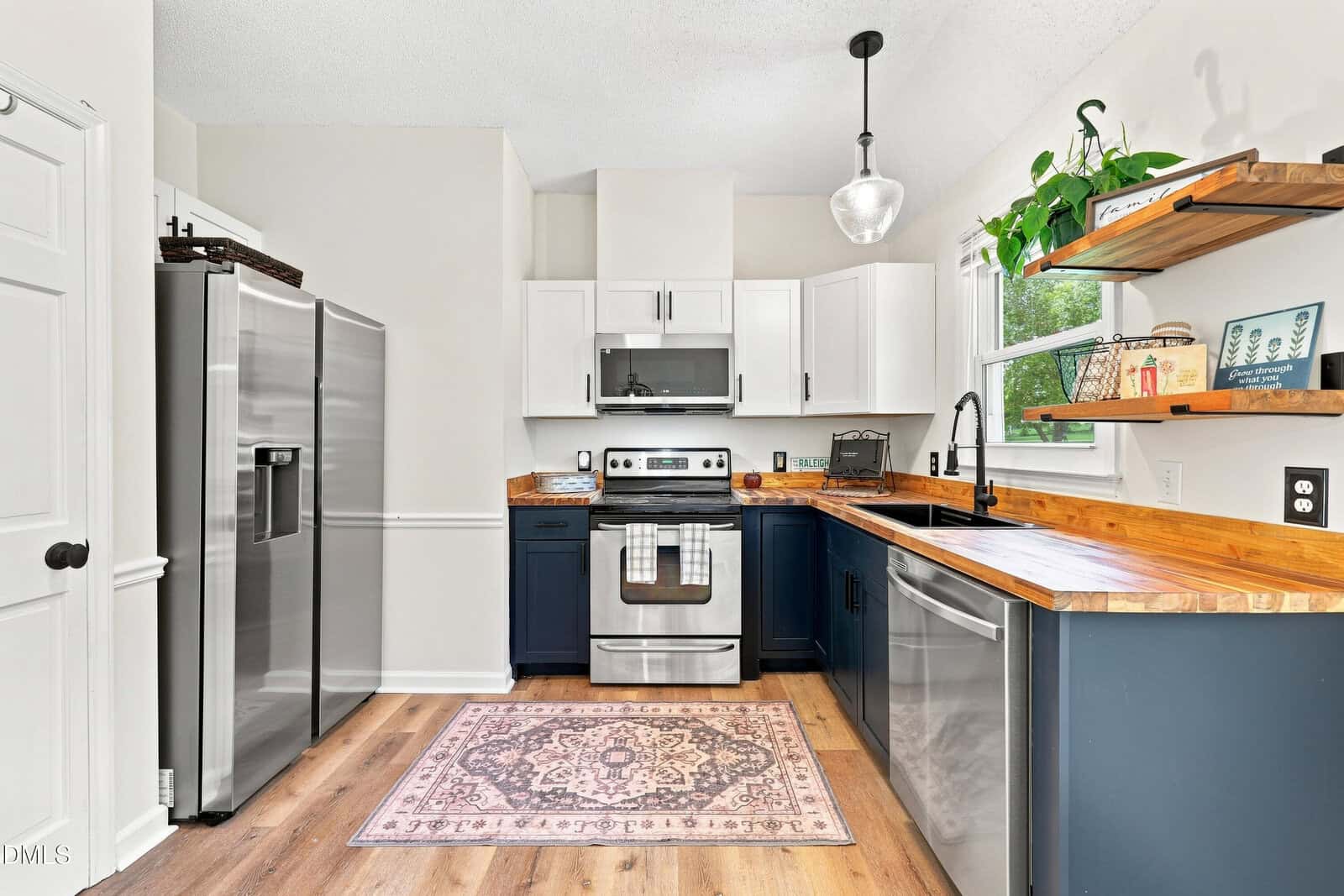 Modern kitchen with stainless steel appliances, a patterned rug, wooden countertops, white upper and navy lower cabinets, open wooden shelves with plants, and a large window providing natural light.