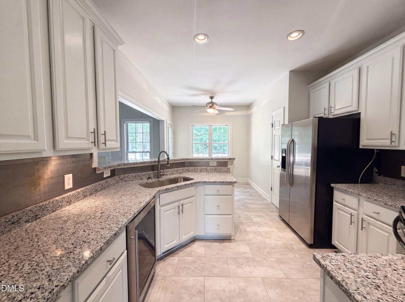Modern kitchen with white cabinets, granite countertops, stainless steel appliances, light tile flooring, under-cabinet lighting, and a ceiling fan in the adjacent dining area near large windows.