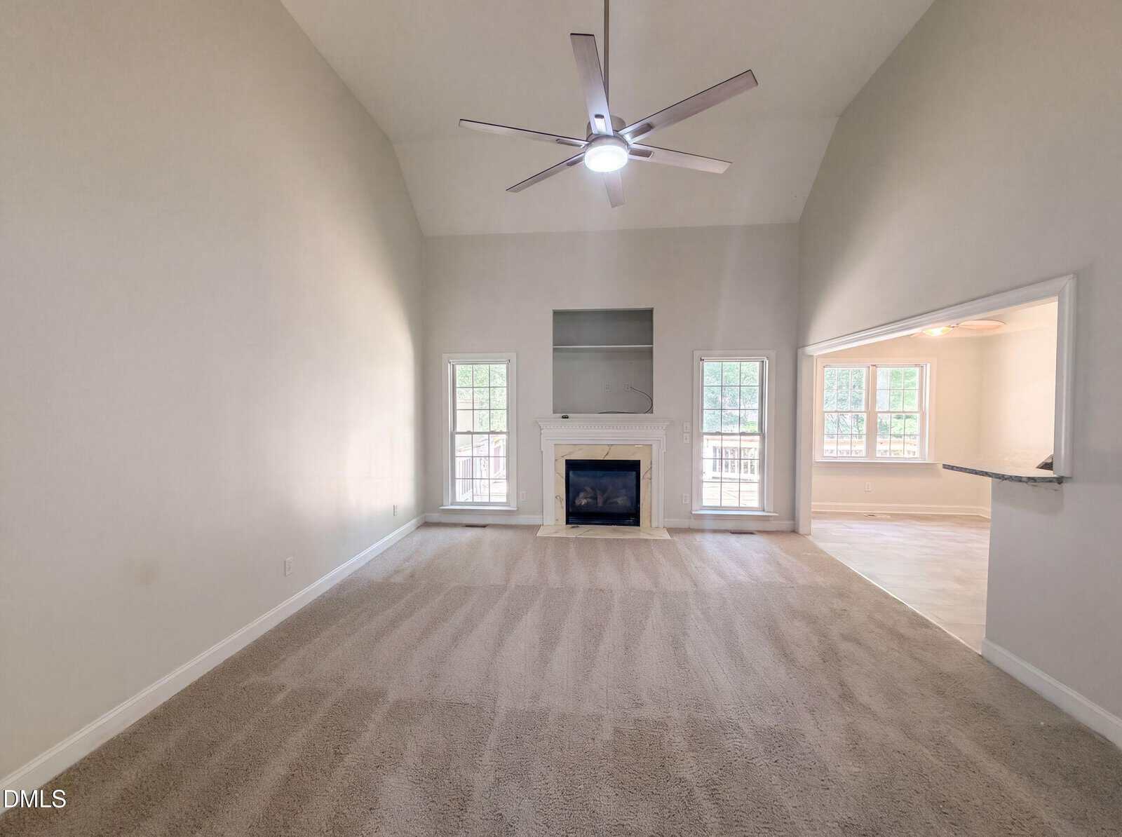 Spacious living room with beige carpet, high ceiling, ceiling fan, fireplace with mantel, two windows on each side, and an open passthrough to an adjacent room. Walls are painted light gray.