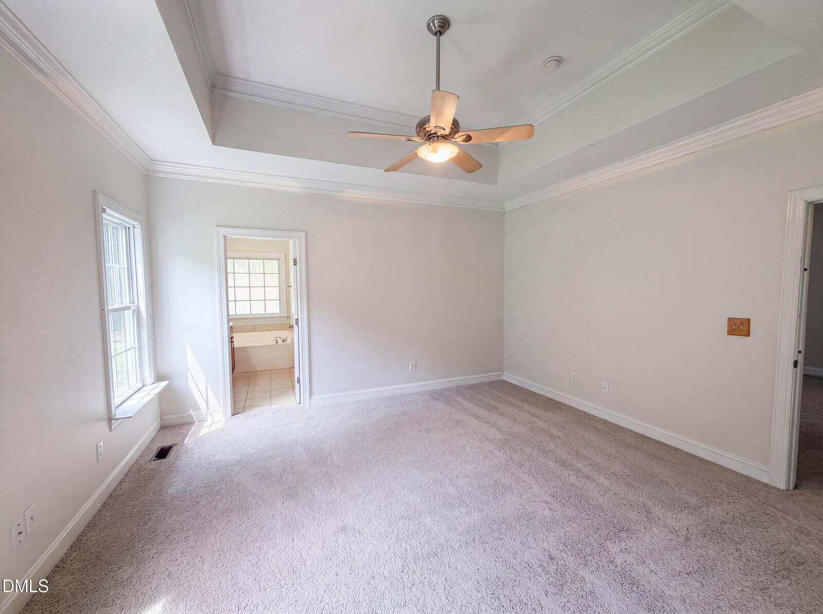 Empty bedroom with beige carpet, cream walls, a ceiling fan with light, tray ceiling detail, and a window. An open door leads to a bathroom with tiled floor and another window visible.