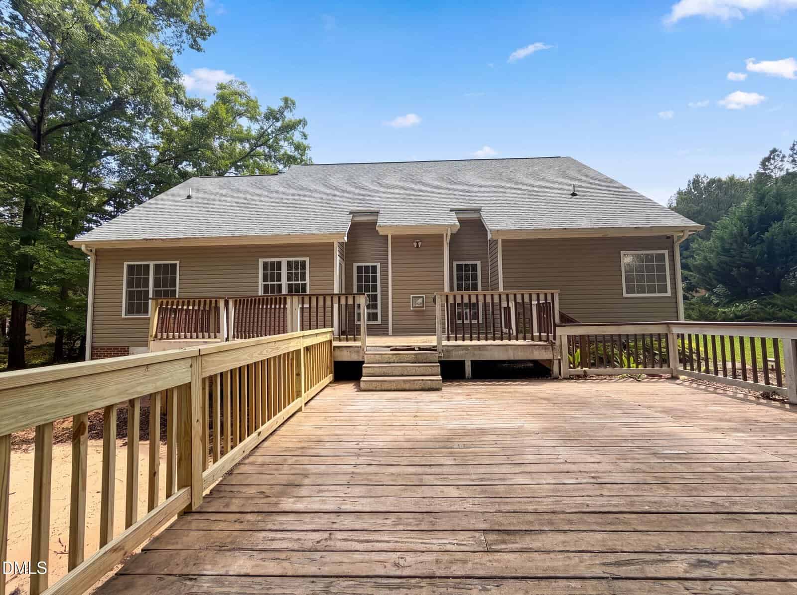 A wide wooden ramp leads up to a tan, single-story house with a gray roof, two small porches, and white-trimmed windows. The house is surrounded by trees under a blue sky with a few clouds.