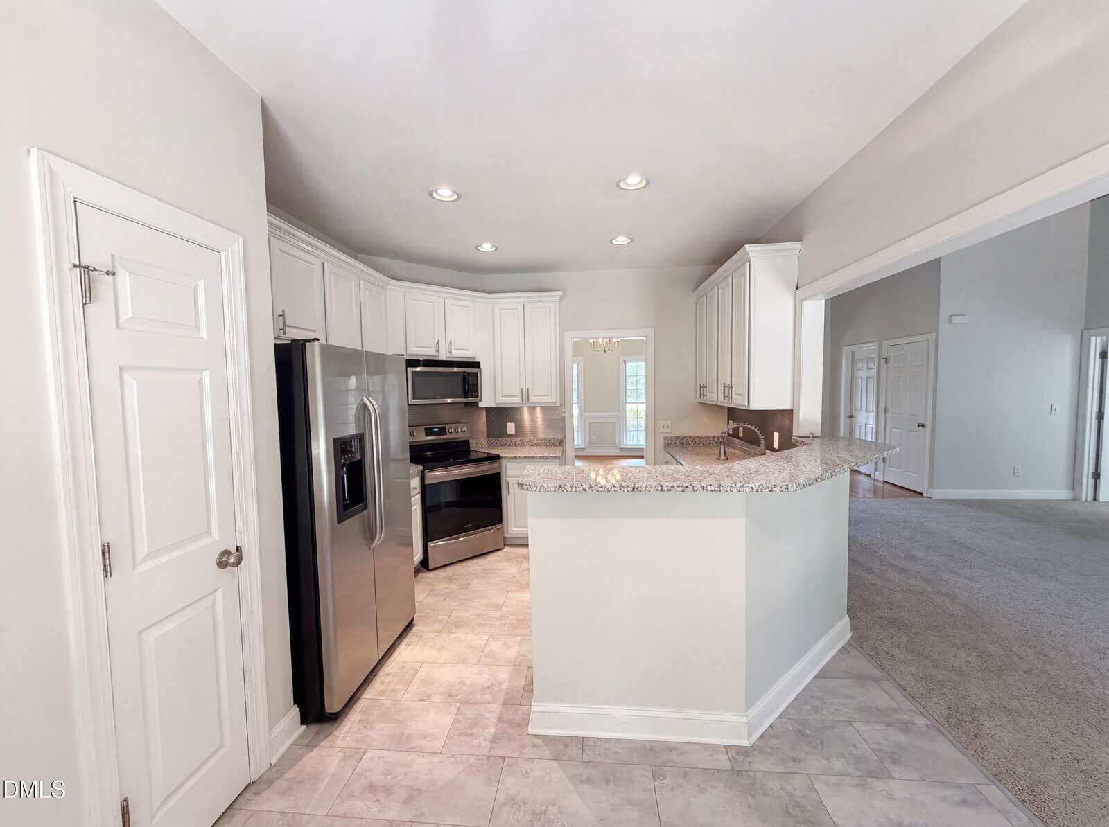 Modern kitchen with stainless steel appliances, white cabinets, granite countertops, tiled floor, and a breakfast bar. The kitchen opens to a carpeted living area with neutral walls and recessed lighting throughout.