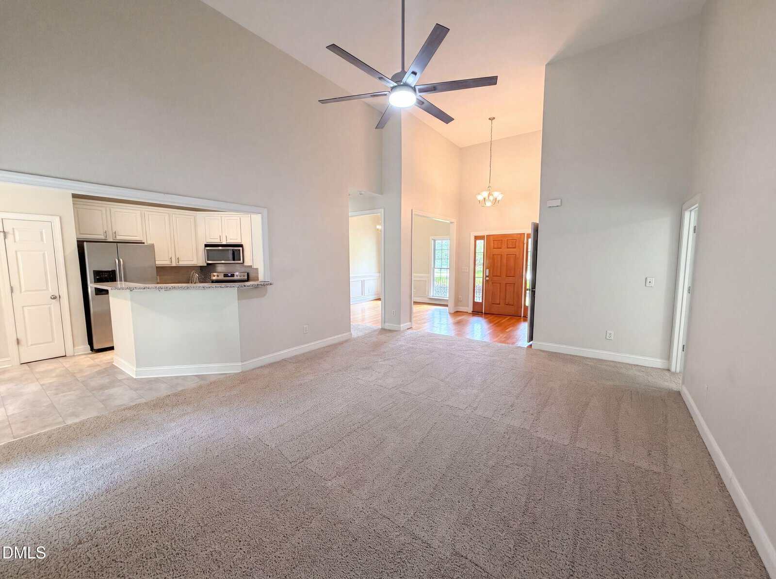 Spacious living room with carpet, high ceiling, ceiling fan, and open kitchen with stainless appliances. Bright entryway with wood flooring and natural light.