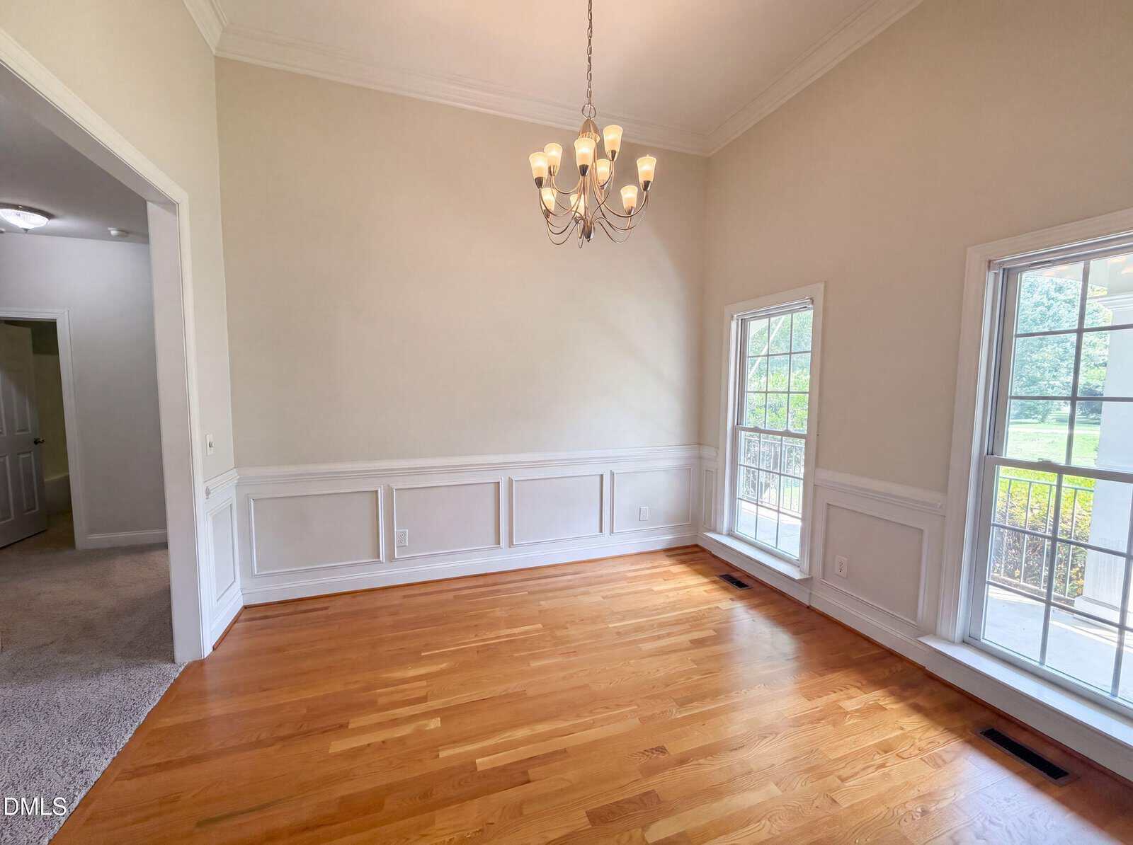 Bright, empty dining room with hardwood floors, a white chandelier, large windows with white trim, beige walls, and wainscoting. An open doorway leads to a carpeted hallway.