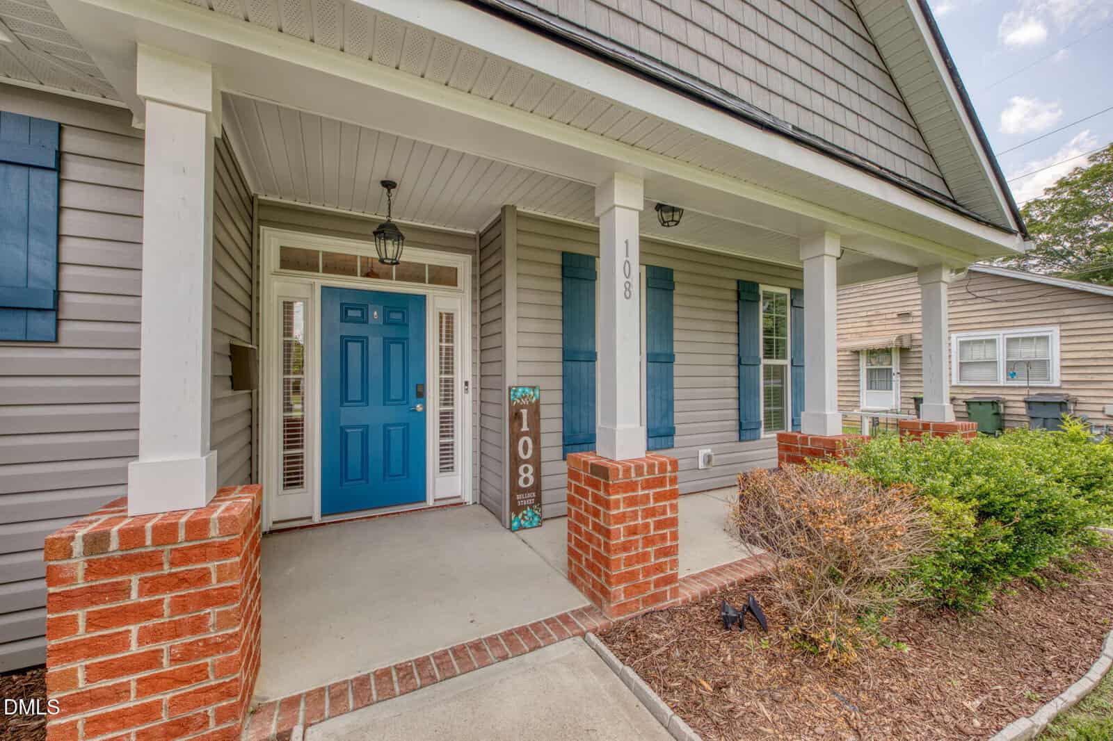 Front porch of a house with a blue door, white pillars, brick accents, gray siding, and a sign displaying the number 108. Small bushes and landscaping are visible near the entryway.