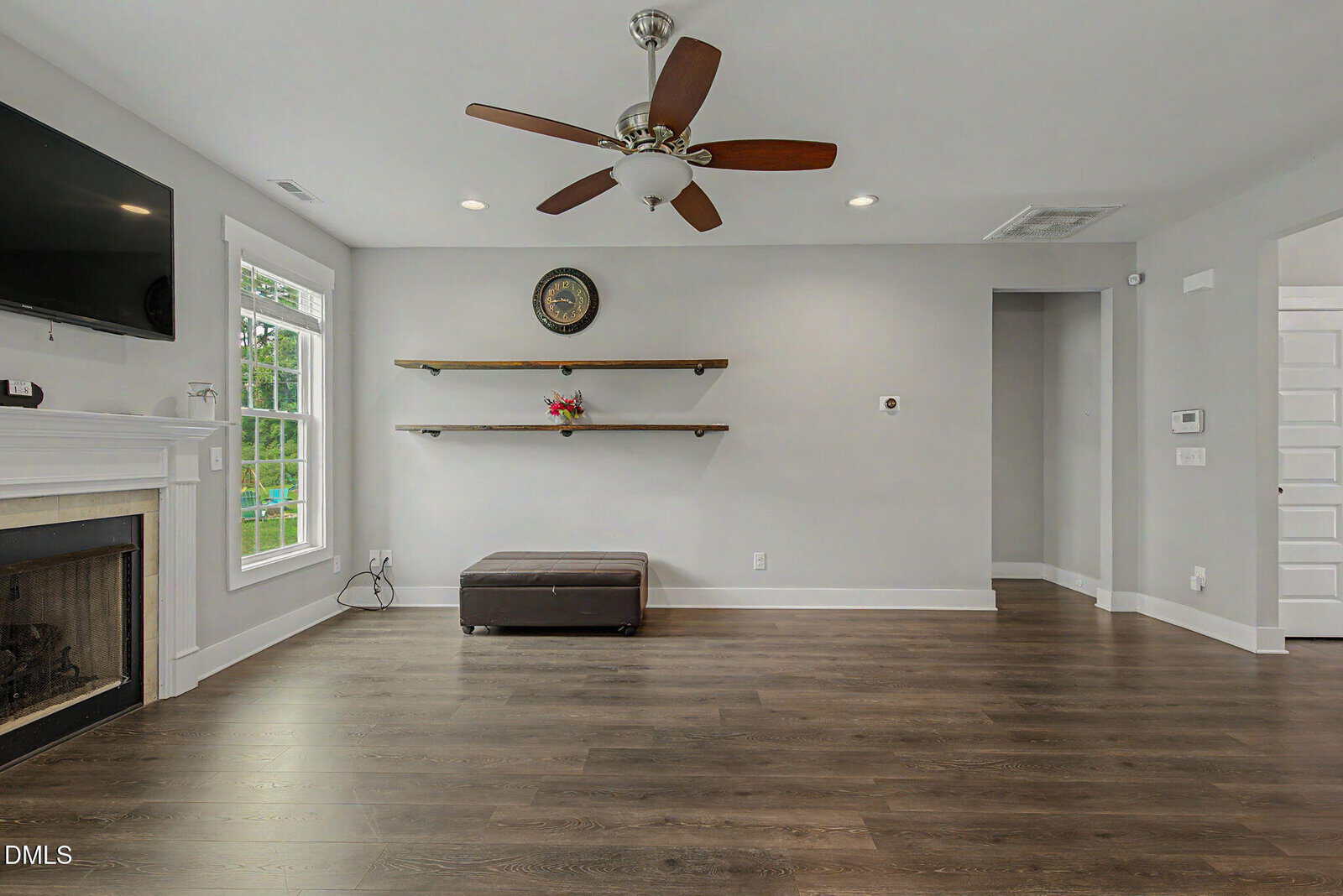 A modern living room with gray walls, wood floors, ceiling fan, and wall-mounted TV. There are two shelves with a small plant and clock on the wall, and a dark ottoman below. A fireplace is on the left side.