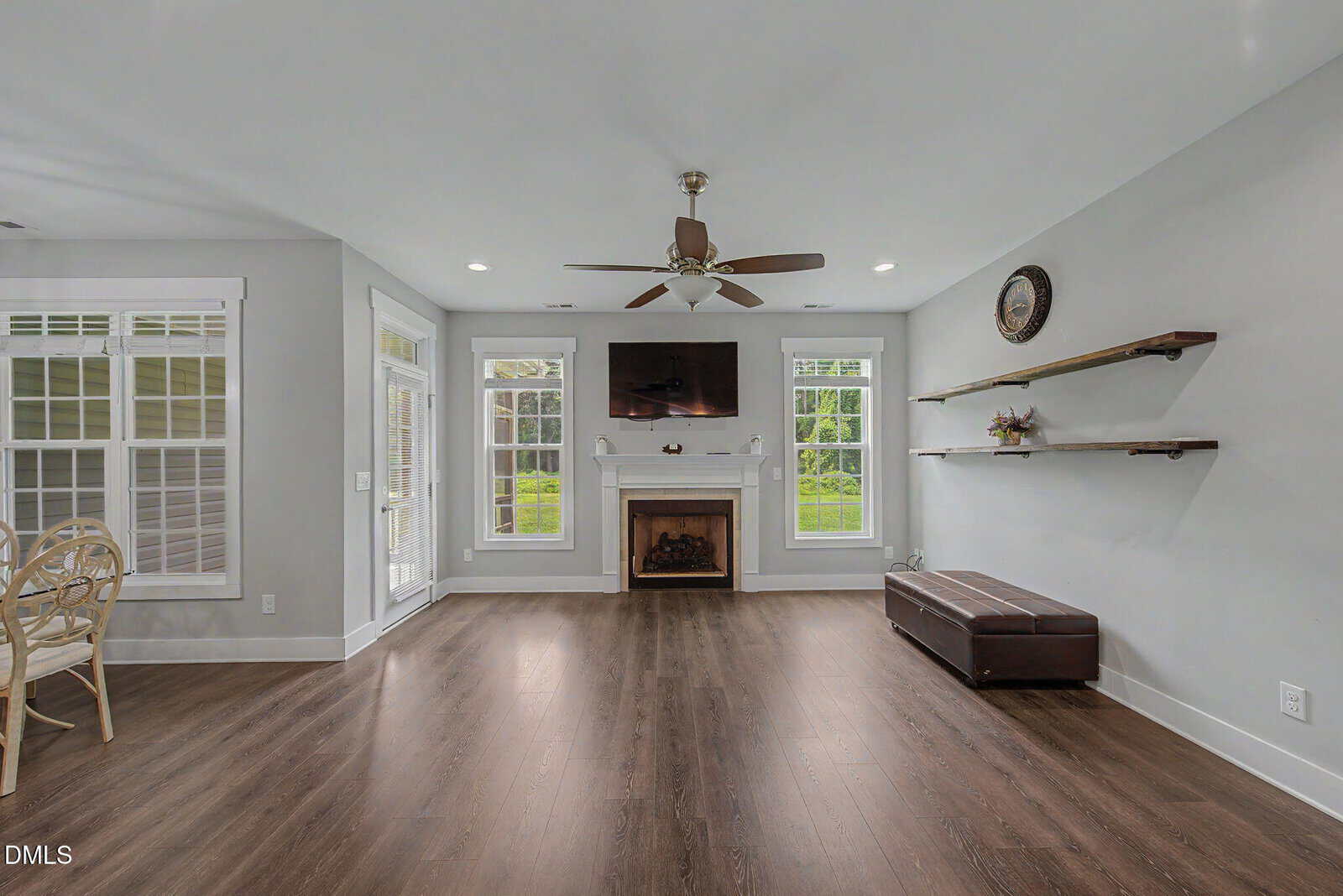 A living room with gray walls, wood flooring, a fireplace below a wall-mounted TV, two floating shelves, a brown bench, a ceiling fan, and large windows letting in natural light.