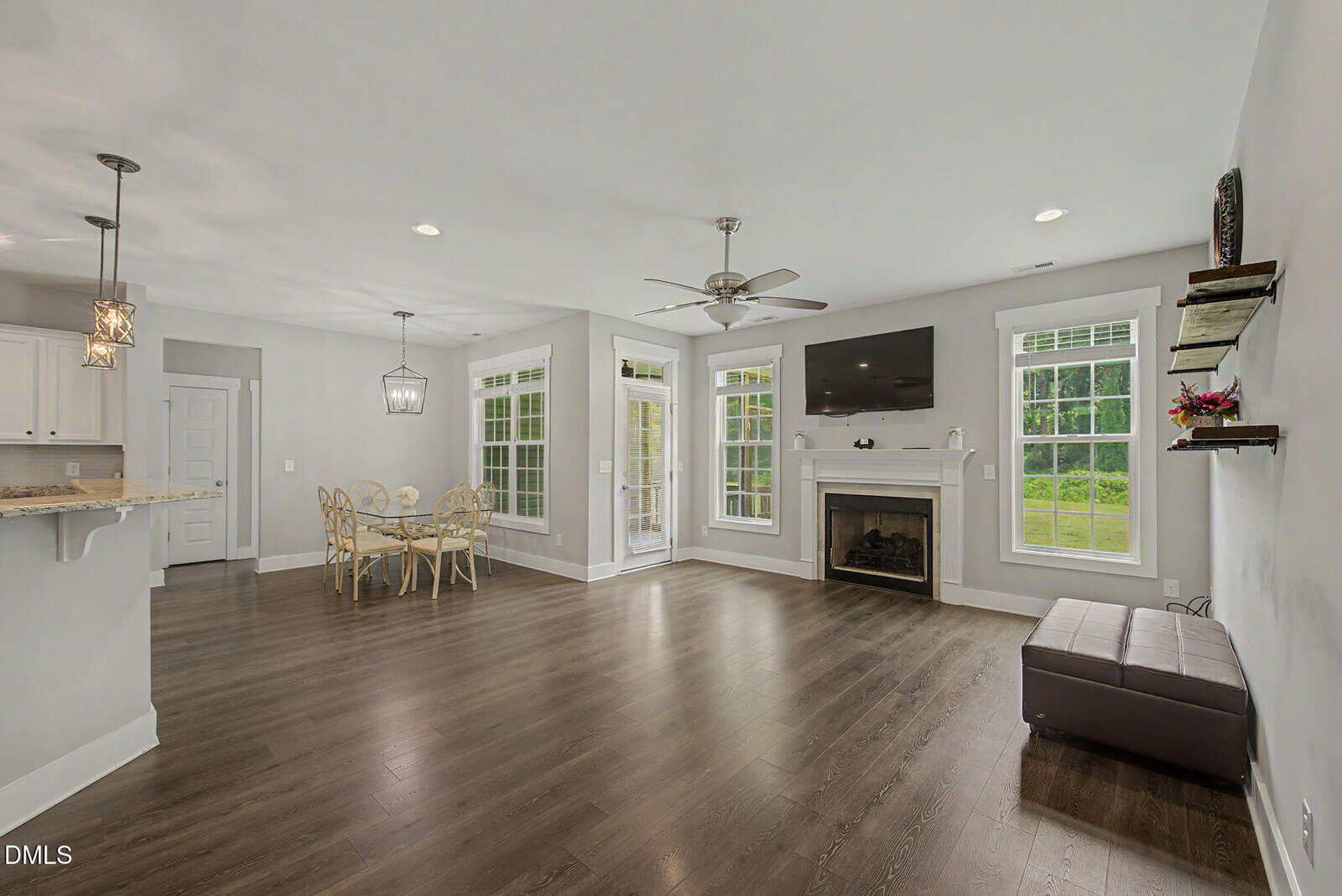 Modern living room with dark wood floors, a fireplace, wall-mounted TV, and large windows. Dining area with a round table and chairs is near the kitchen, which features pendant lights and a breakfast bar.