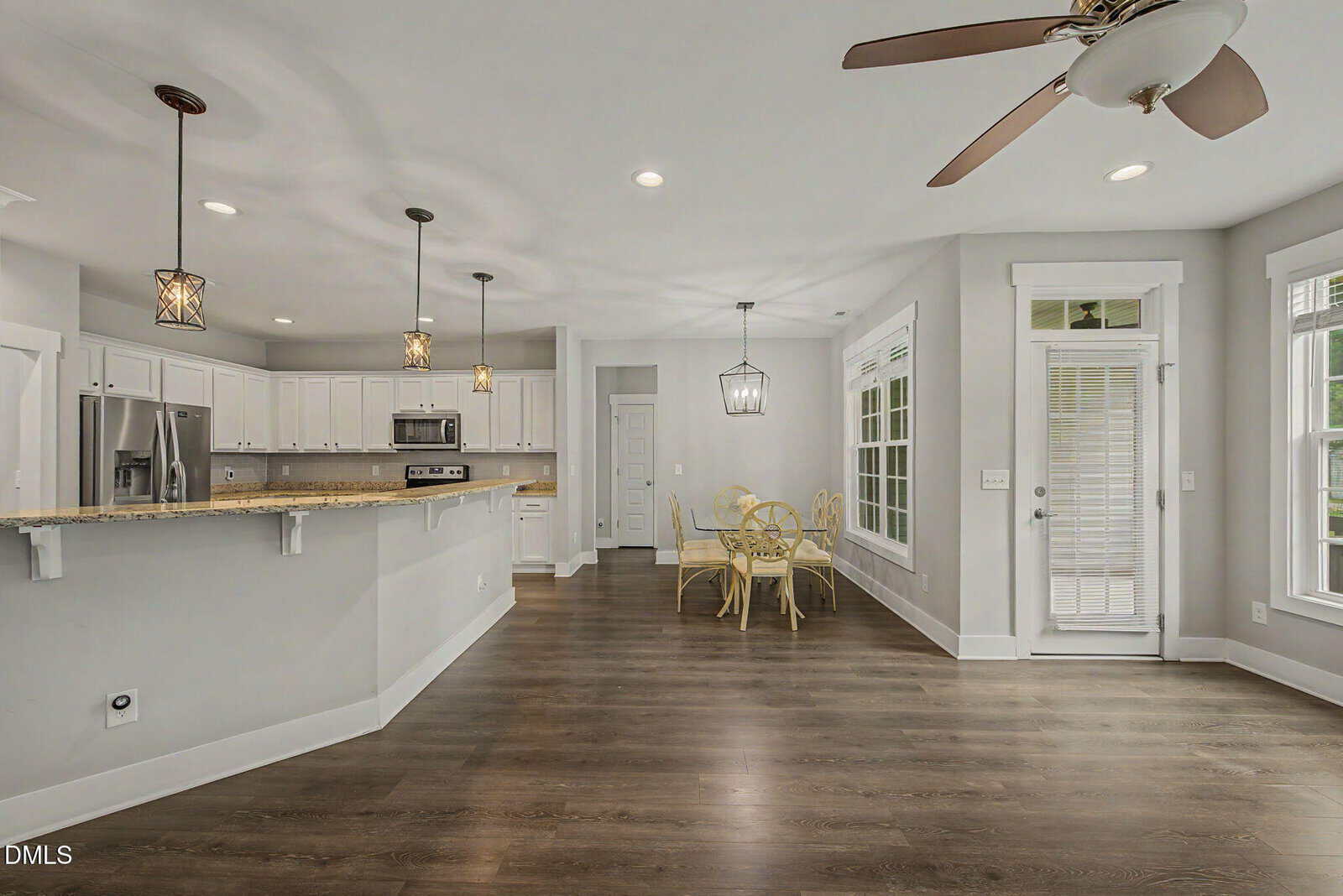 Modern open-plan kitchen and dining area with wood flooring, white cabinets, stainless steel appliances, pendant lights, and large windows letting in ample natural light.