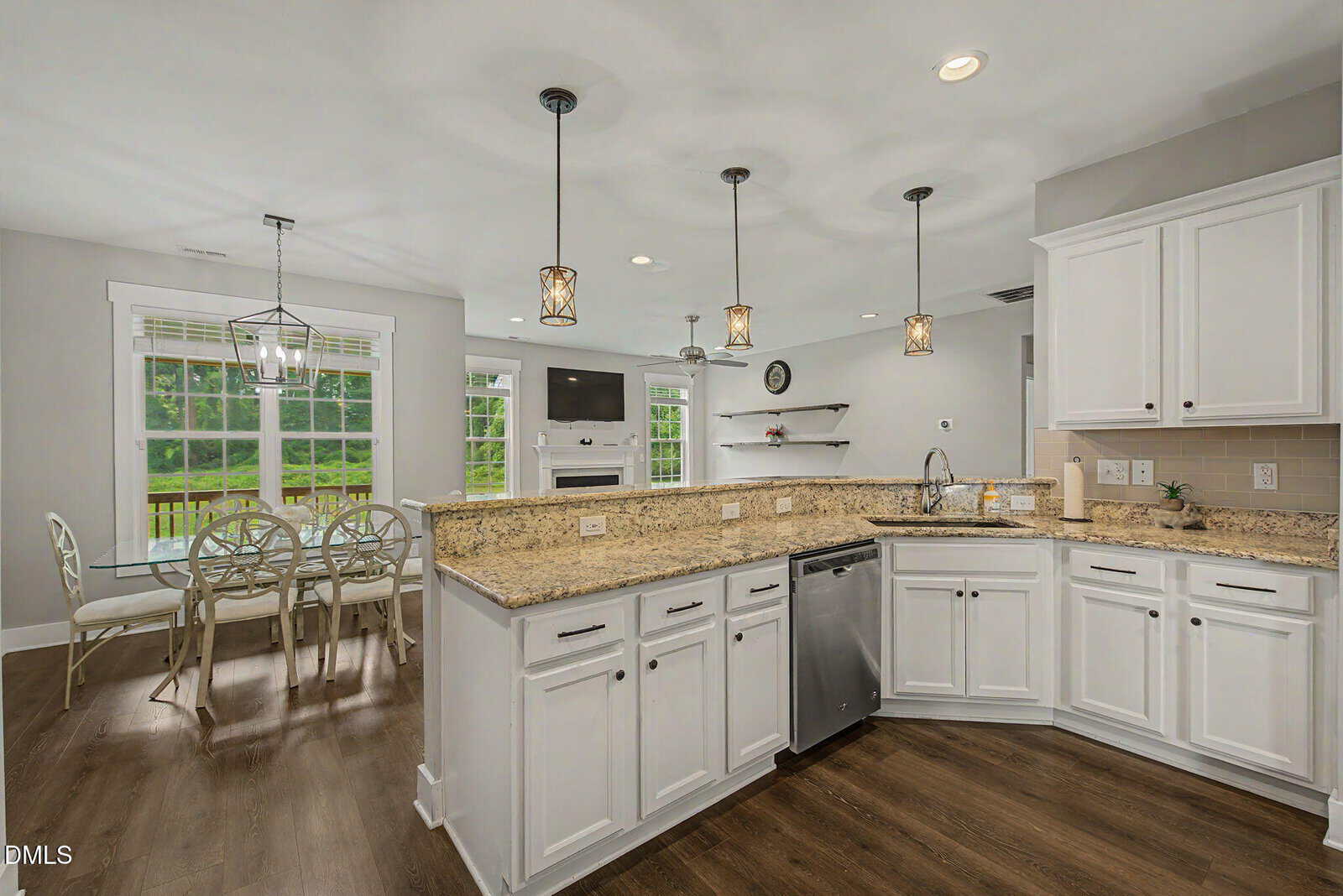 Modern kitchen with white cabinets, granite countertops, stainless steel appliances, pendant lights, and open shelves. Large windows and a dining area with a round table are visible in the background.