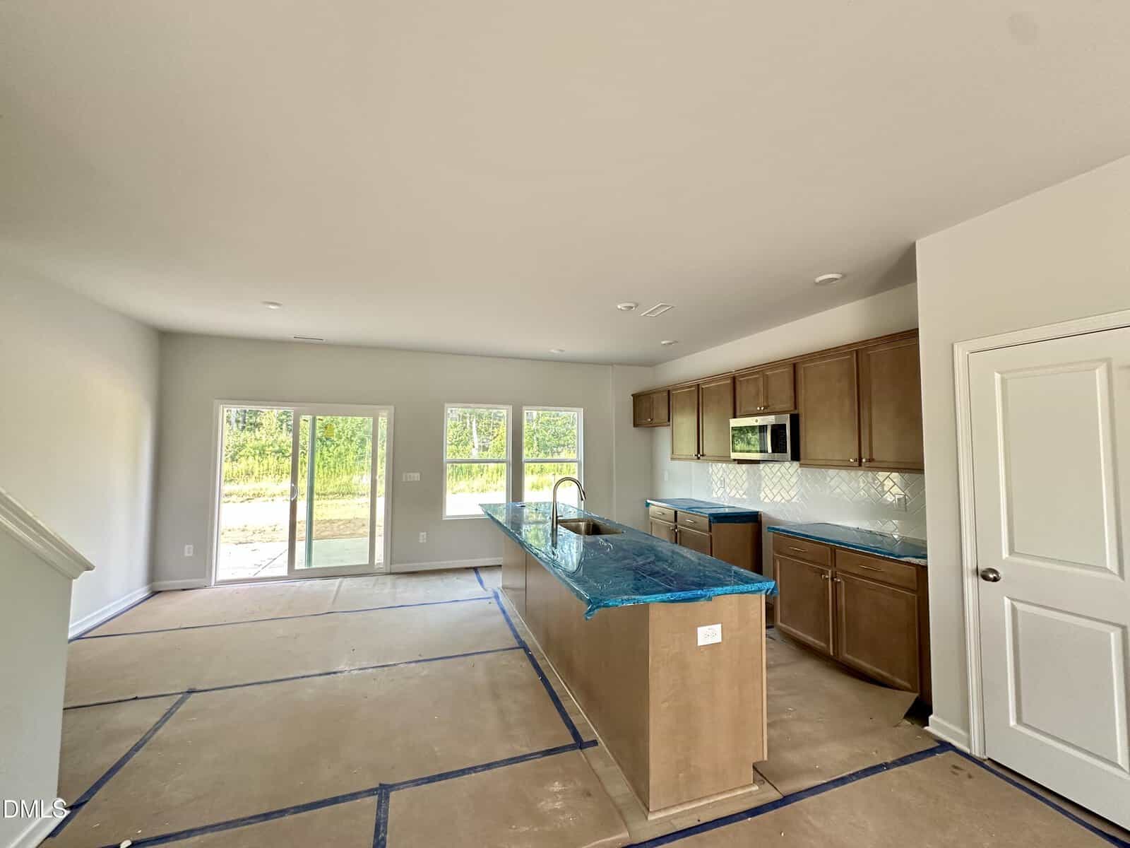 New kitchen with brown cabinets, large island with blue protective wrap, white backsplash, and stainless steel appliances. Unfinished floors are covered for protection. Large windows and a glass door let in natural light.