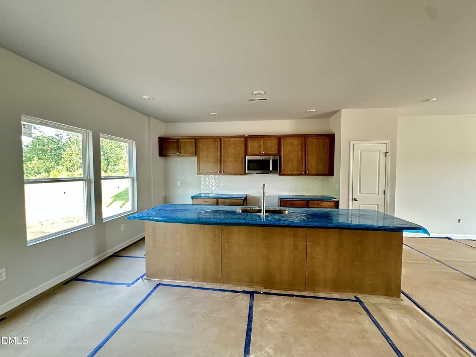 A modern kitchen with wood cabinets, a large island with a blue countertop, stainless steel appliances, and large windows letting in natural light. The floor is covered for protection during construction.