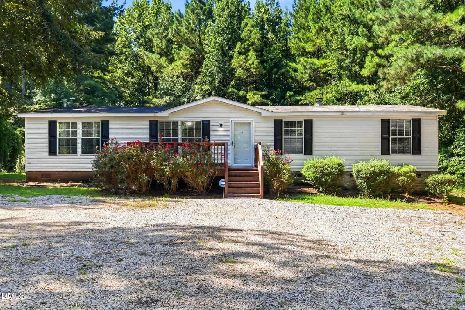 A white, single-story house with black shutters, a small front porch, wooden steps, surrounding bushes, and a gravel driveway sits in front of a background of tall green trees at 115 Carter Lane in Warrenton.