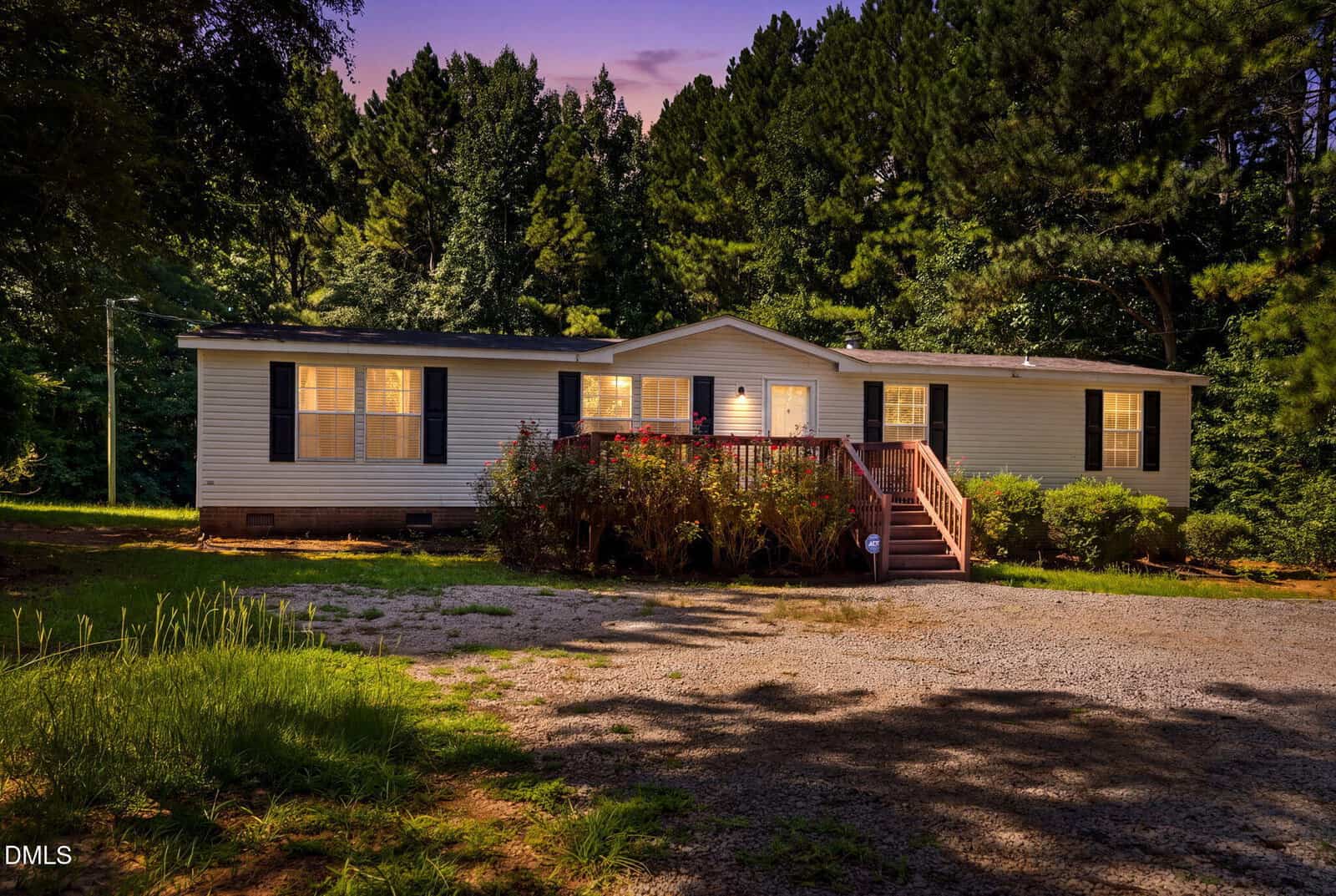 A single-story house with lit windows and a front deck sits surrounded by trees at dusk, with a gravel driveway and landscaped shrubs in the foreground.